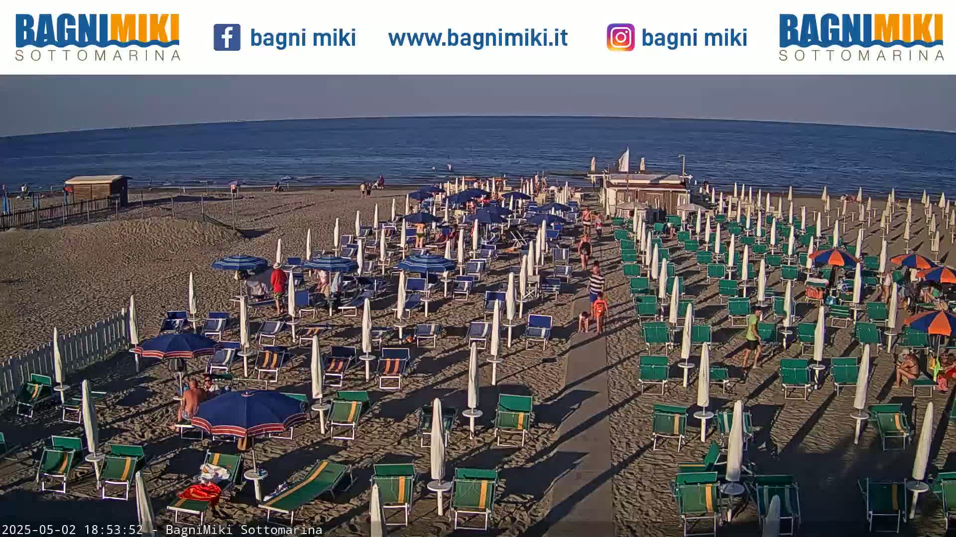 A sandy beach with many beach umbrellas and lounge chairs is crowded with people on a sunny day, and a calm ocean is visible in the background.
