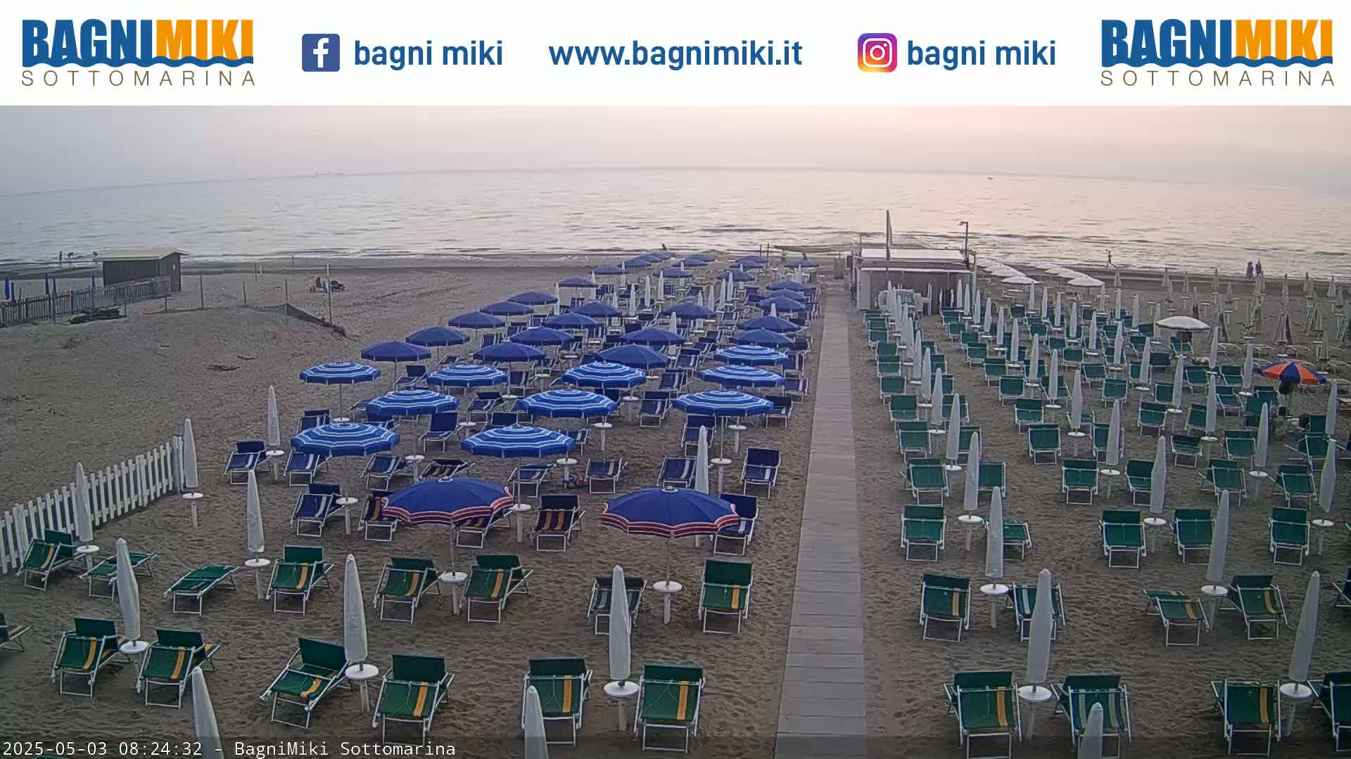 A mostly sunny beach with rows of closed umbrellas and lounge chairs, overlooking a calm sea.