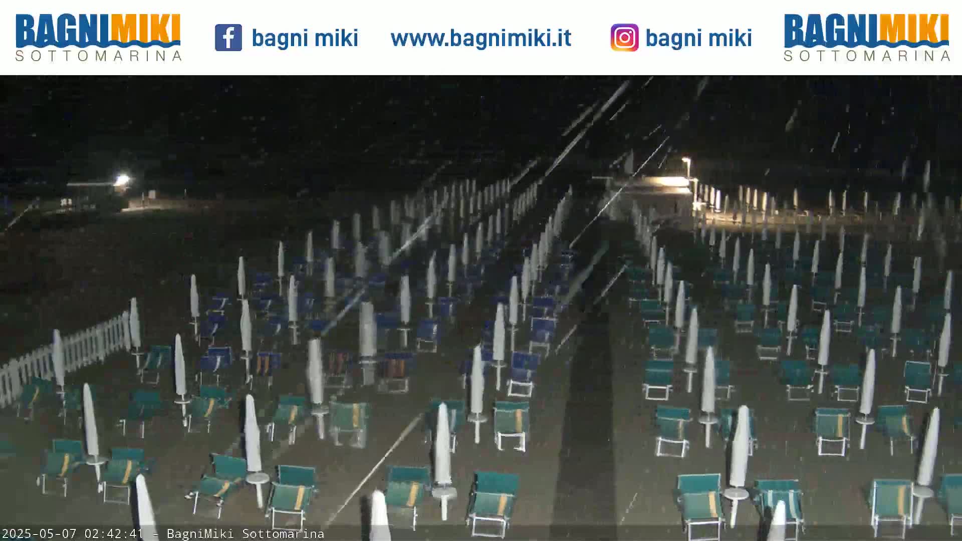 Rows of beach umbrellas and chairs sit on a dark, sandy beach at night under a light rain.