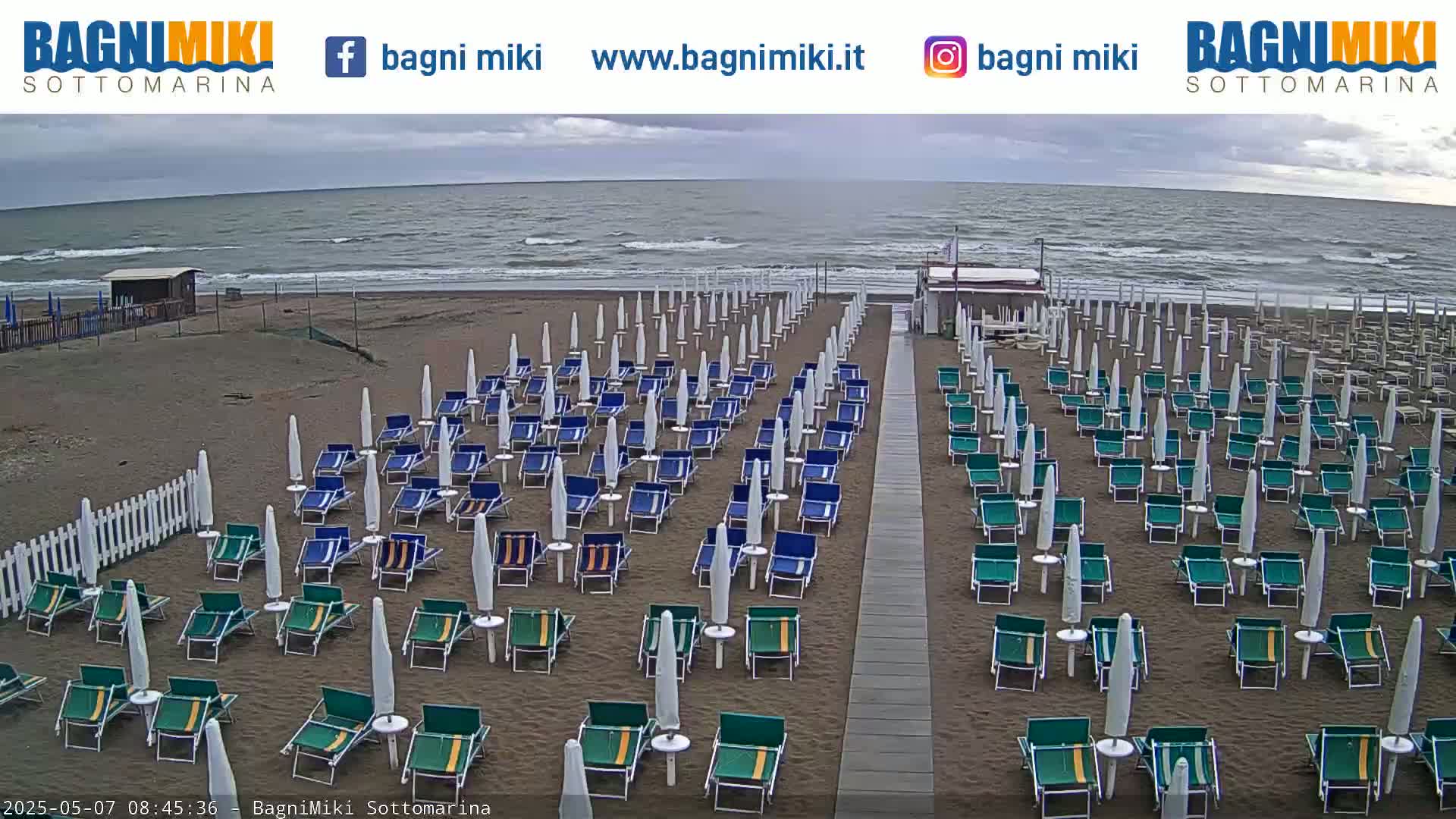 Rows of beach chairs and umbrellas sit on a sandy beach under an overcast sky, with a gray sea visible in the background.