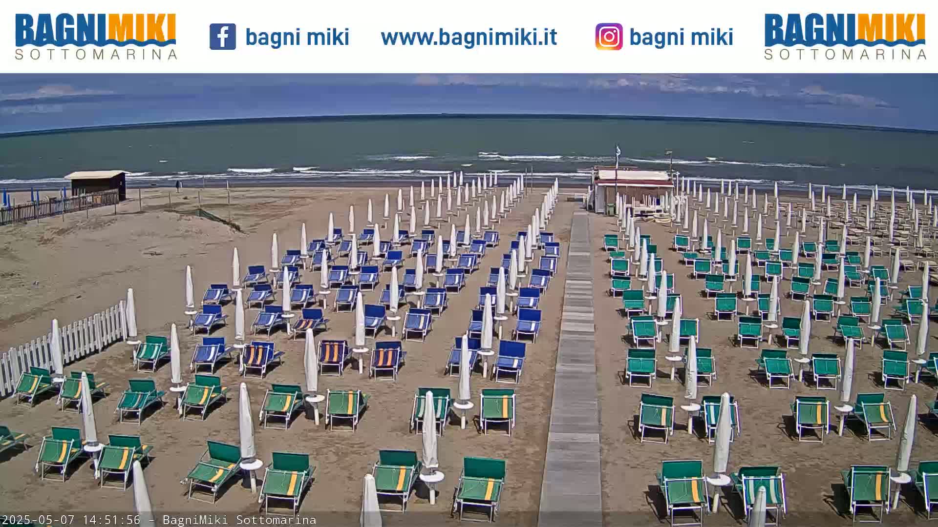 A sandy beach with numerous beach umbrellas and lounge chairs arranged in rows under a mostly sunny sky, overlooking a calm sea.