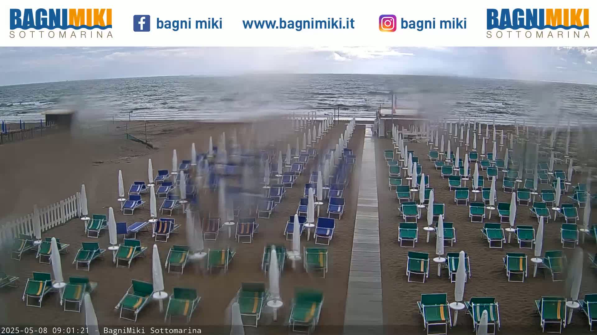 Rows of beach chairs and umbrellas sit empty on a sandy beach under a cloudy, possibly rainy sky, with a choppy sea in the background.