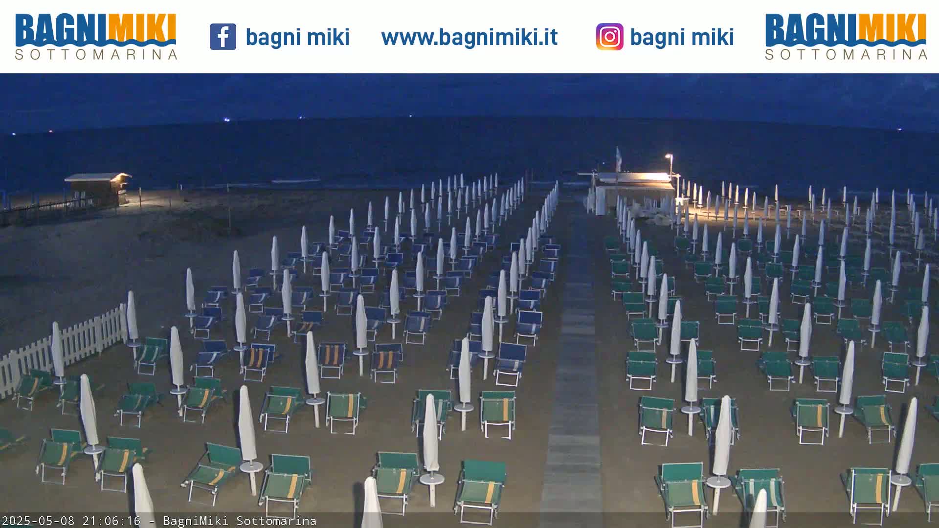 Rows of beach umbrellas and chairs stand on a sandy beach at night, next to a calm, dark ocean.