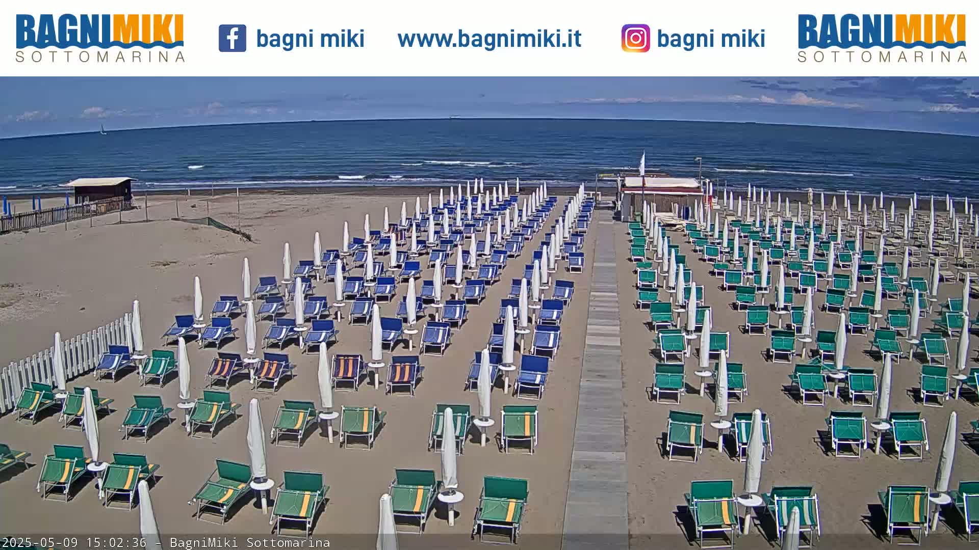 A sandy beach with numerous rows of beach umbrellas and lounge chairs is shown, under a mostly clear sky and calm ocean waves.