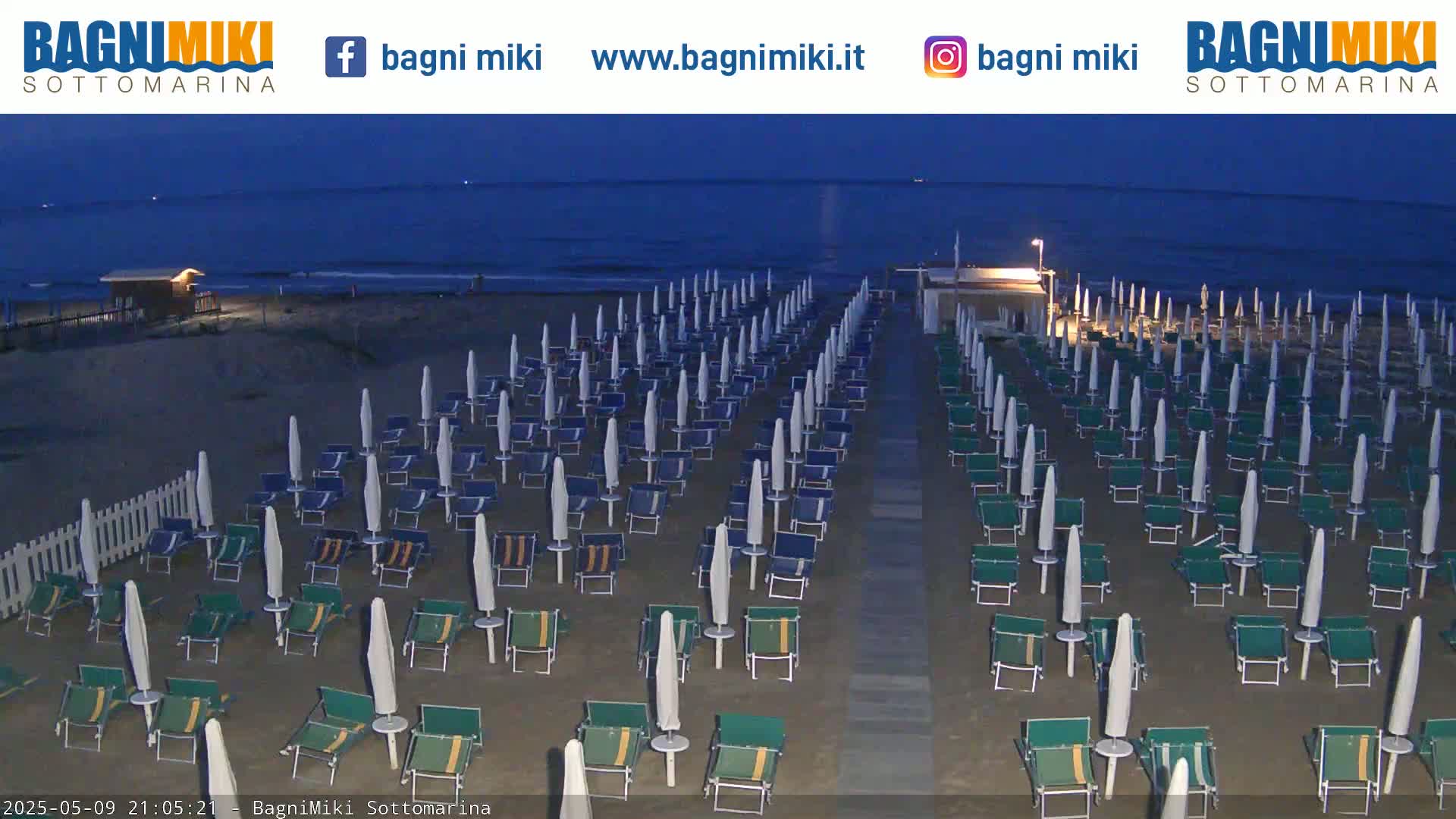 Rows of closed beach umbrellas and lounge chairs sit on a dark sandy beach at twilight, next to a calm, dark blue ocean.