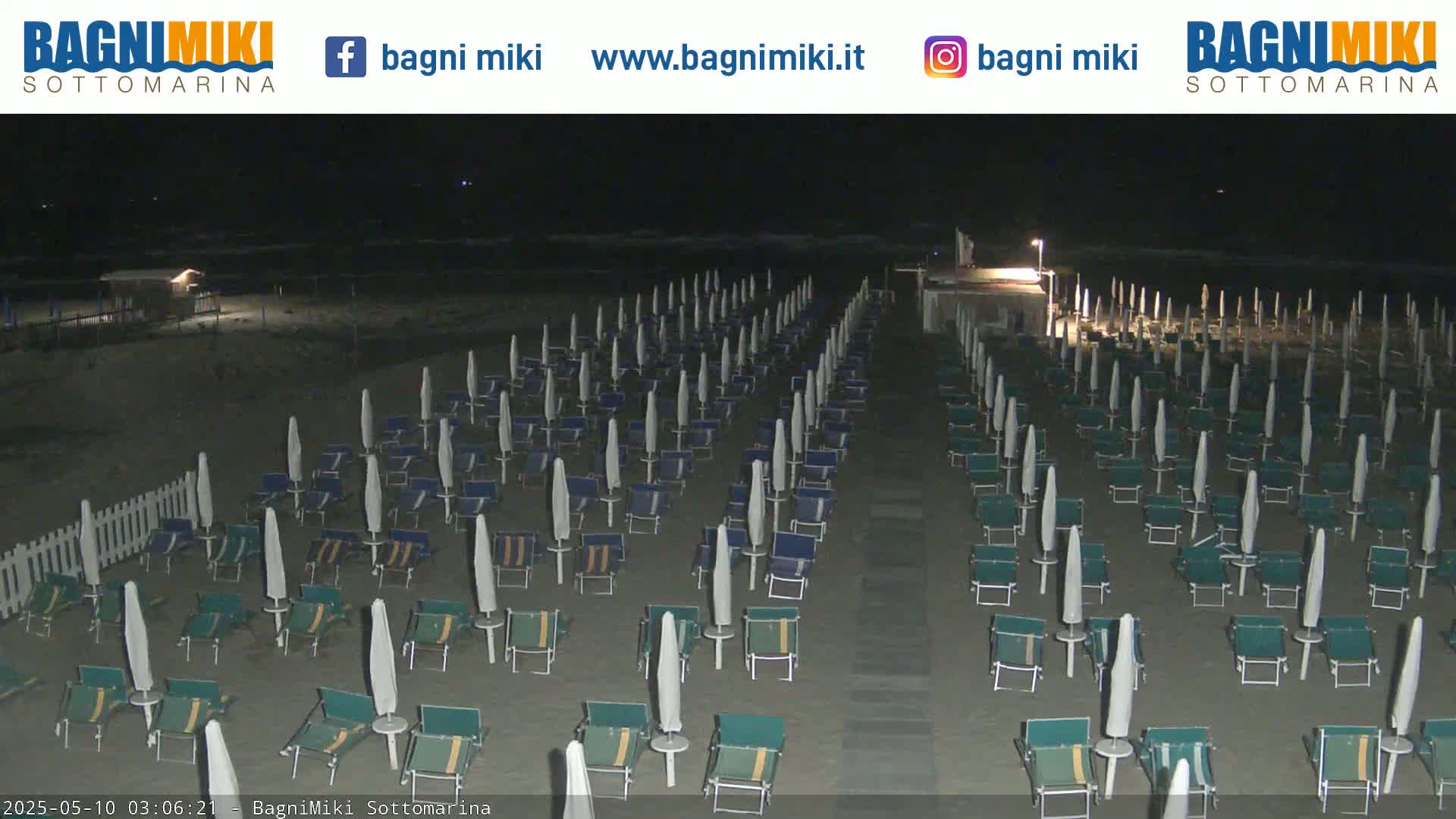 Rows of closed beach umbrellas and lounge chairs stand on a dark, sandy beach at night.