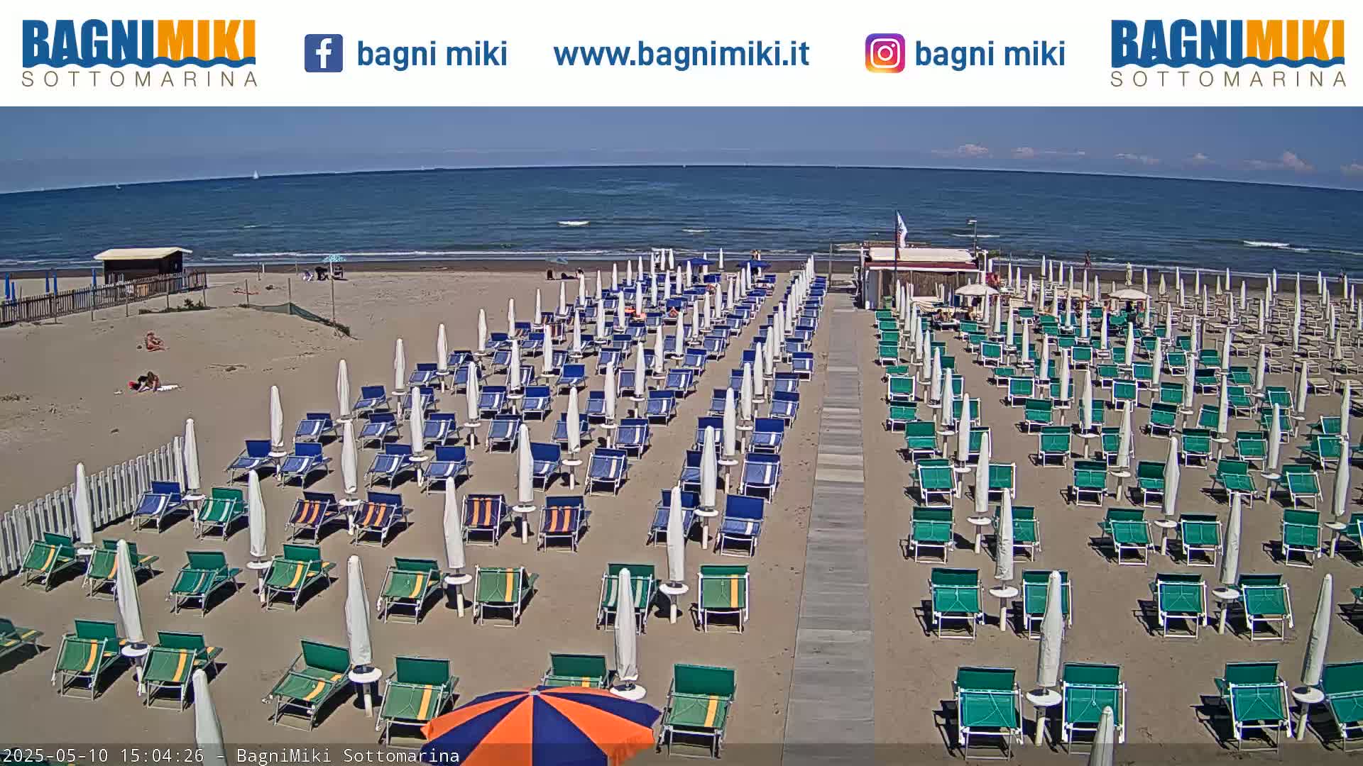 Rows of beach umbrellas and lounge chairs line a sandy beach next to a calm ocean under a sunny sky.