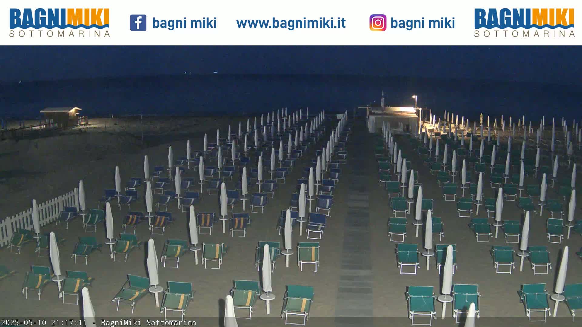 Rows of closed beach umbrellas and chairs sit on a sandy beach at night under a dark sky.