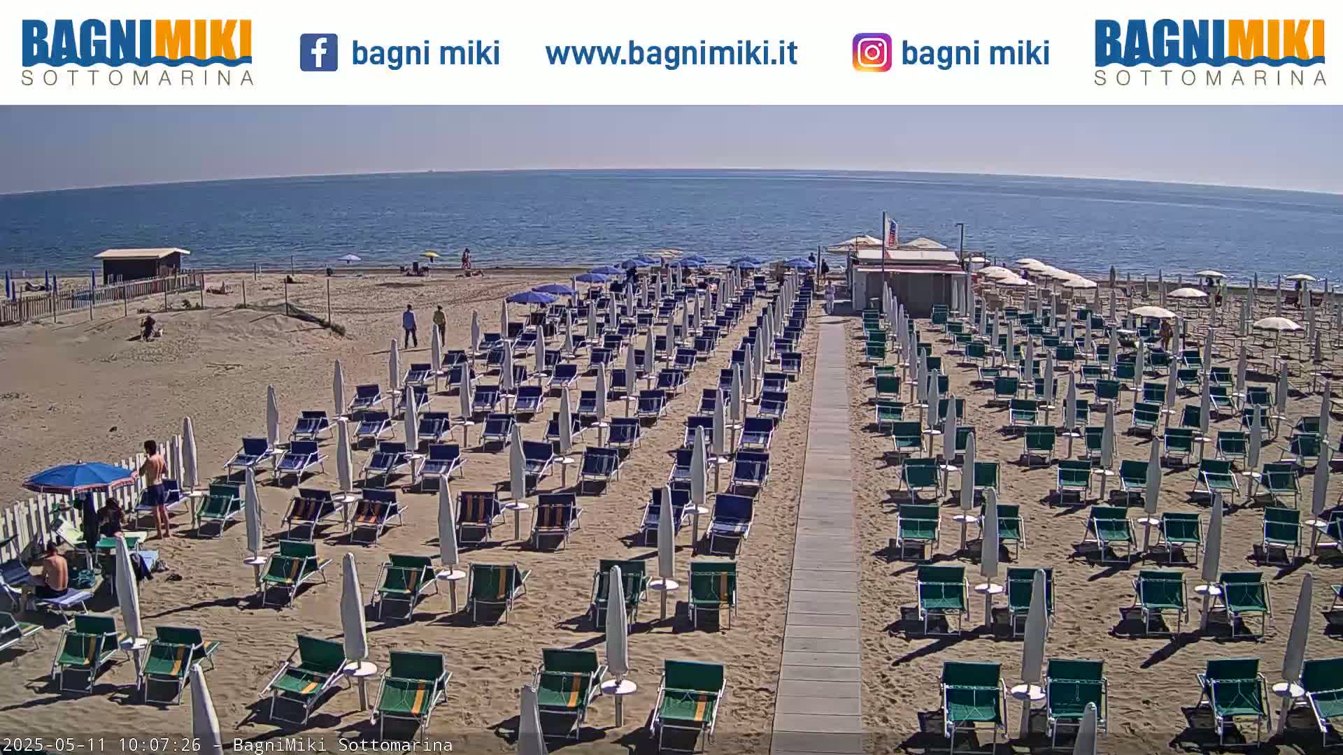 A sandy beach with numerous rows of beach umbrellas and lounge chairs is situated next to a calm ocean under a sunny sky.