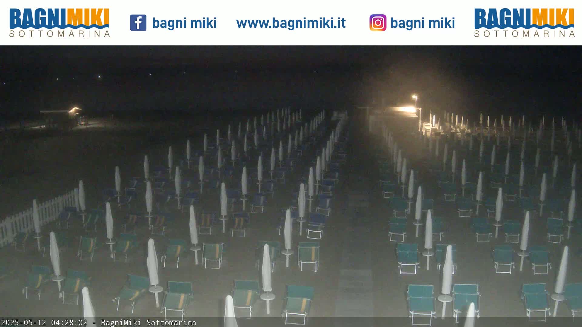 A dark beach at night is filled with rows of closed beach umbrellas and chairs under a hazy sky.