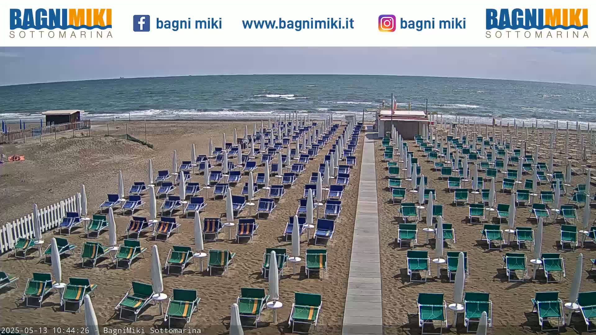 A sandy beach with numerous beach umbrellas and chairs lines a walkway leading to a calm, somewhat wavy ocean under a bright, sunny sky.