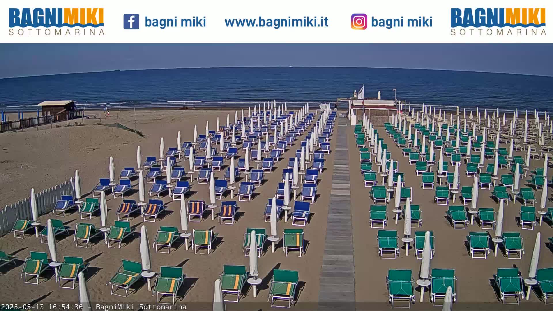A sandy beach with numerous rows of green lounge chairs and white umbrellas under a clear sky and calm ocean.