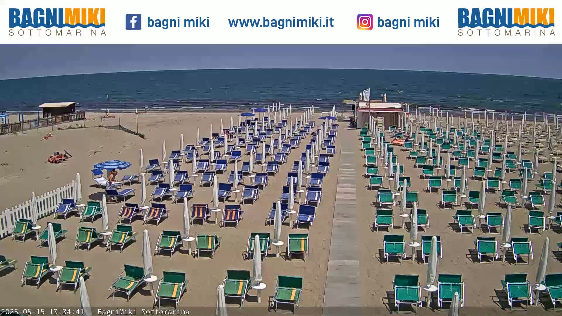 A sandy beach with numerous beach umbrellas and lounge chairs is visible under a sunny sky, next to a calm ocean.
