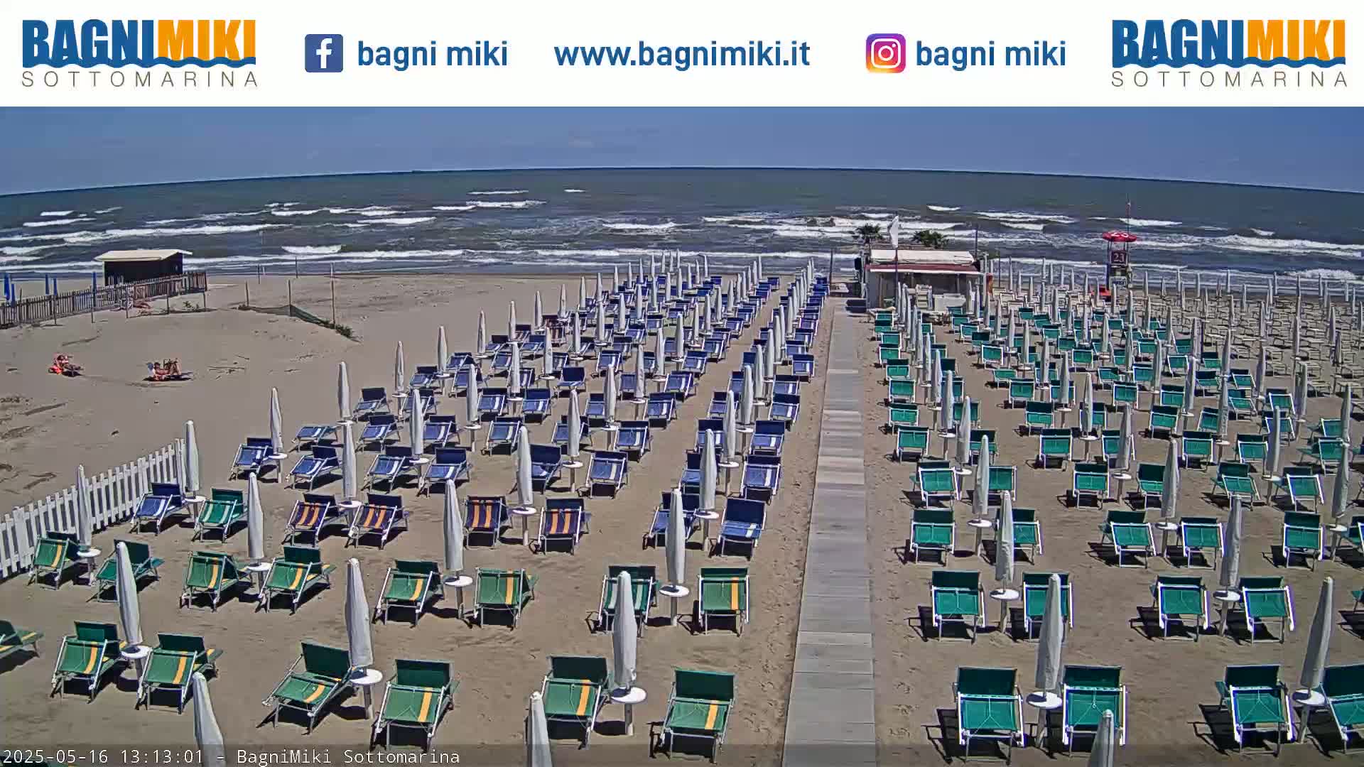 Rows of beach umbrellas and chairs line a sandy beach next to a wavy ocean under a bright sunny sky.