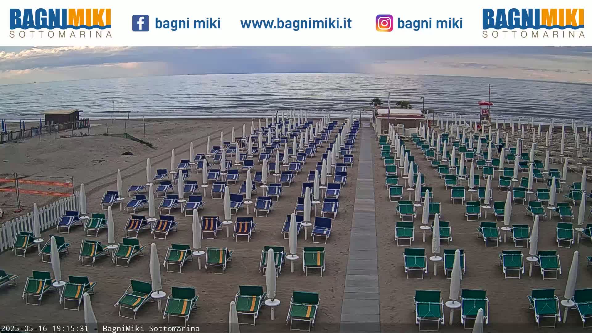 Rows of beach chairs and umbrellas sit on a sandy beach next to a calm ocean under an overcast sky.