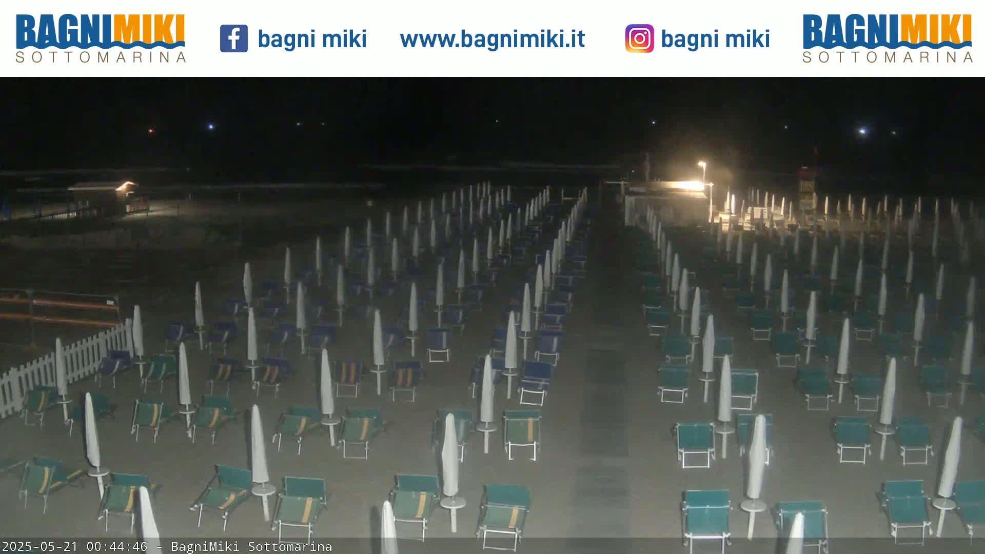 A nighttime beach scene shows numerous closed beach umbrellas and lounge chairs arranged in neat rows on a sandy beach under dark skies.