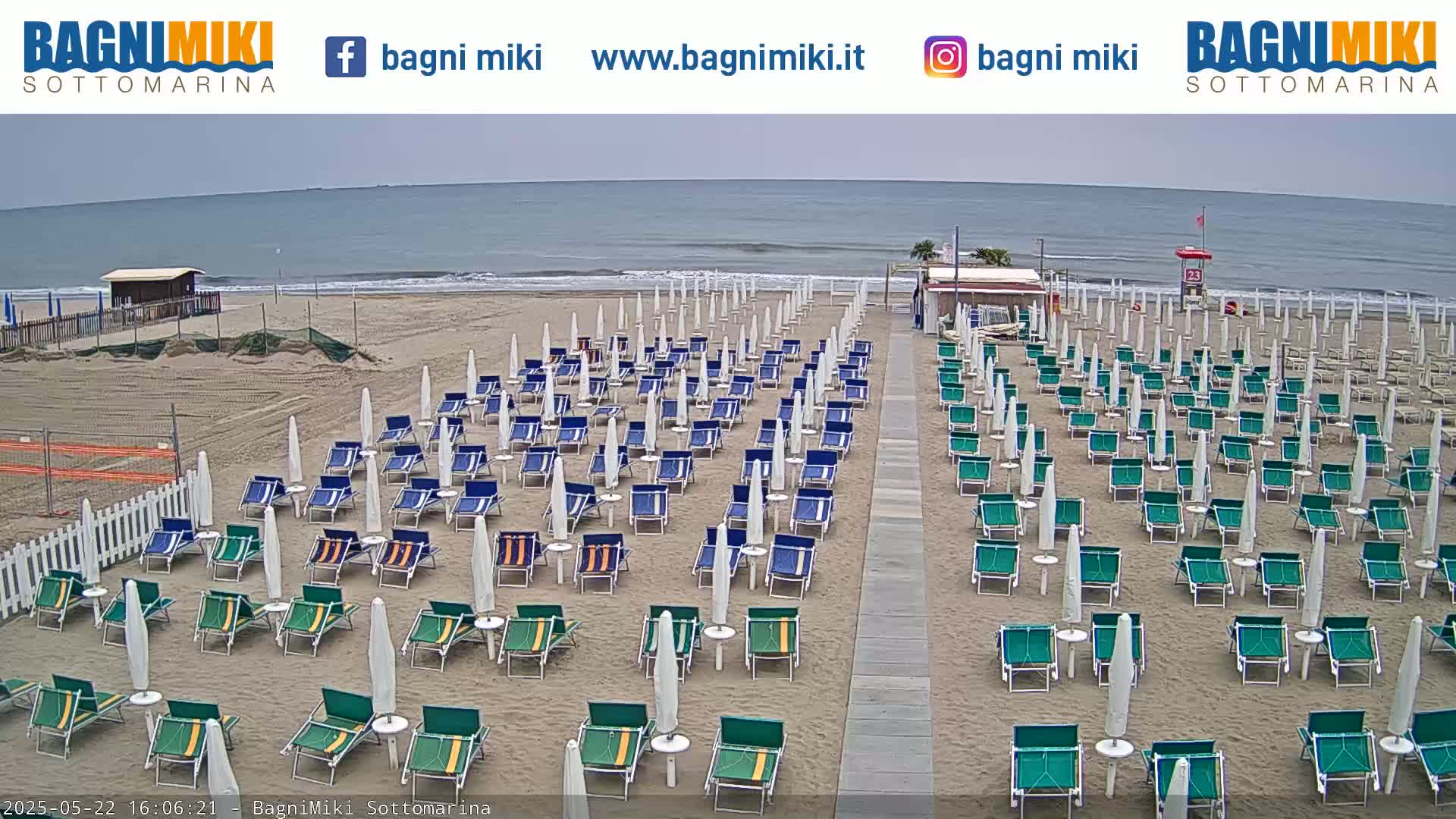 A sandy beach with numerous rows of closed beach umbrellas and chairs under an overcast sky.