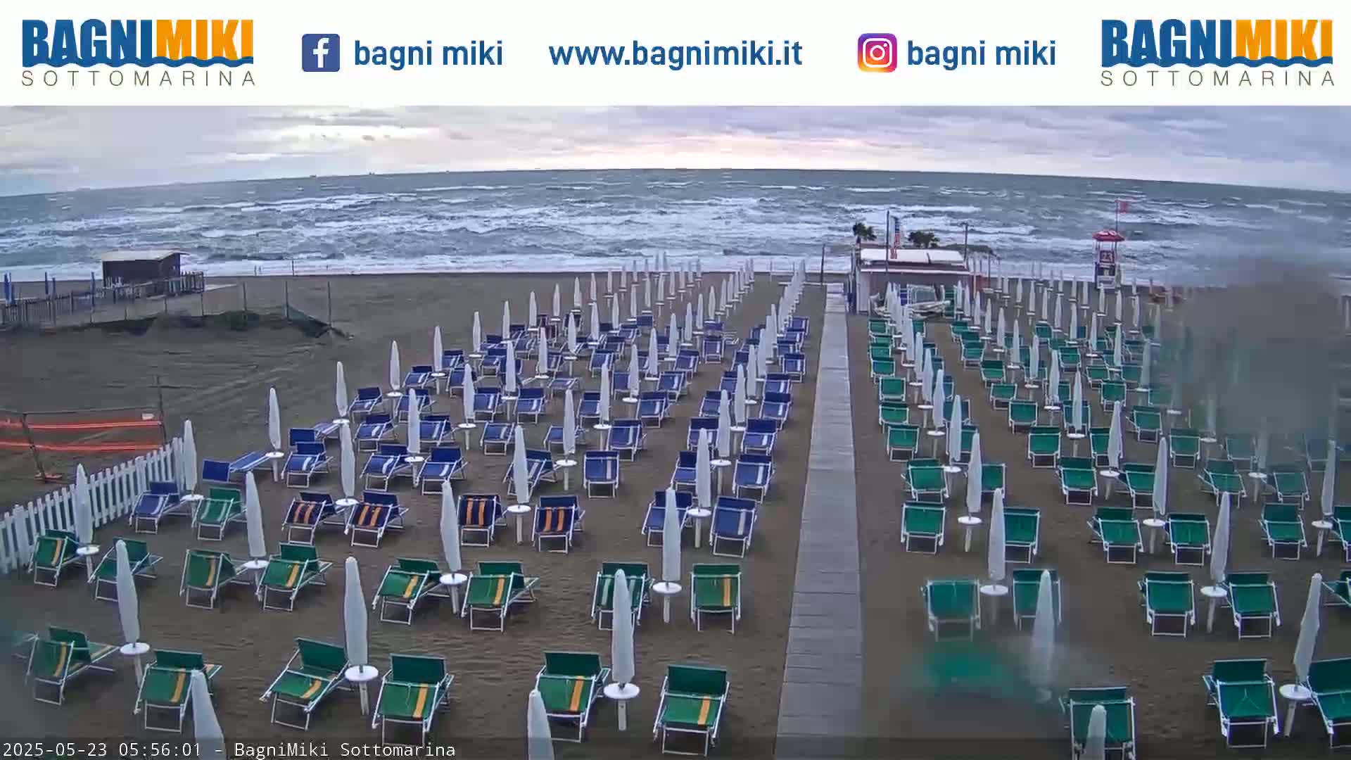 Rows of beach chairs and umbrellas sit on a sandy beach under an overcast sky, with a choppy sea in the background.