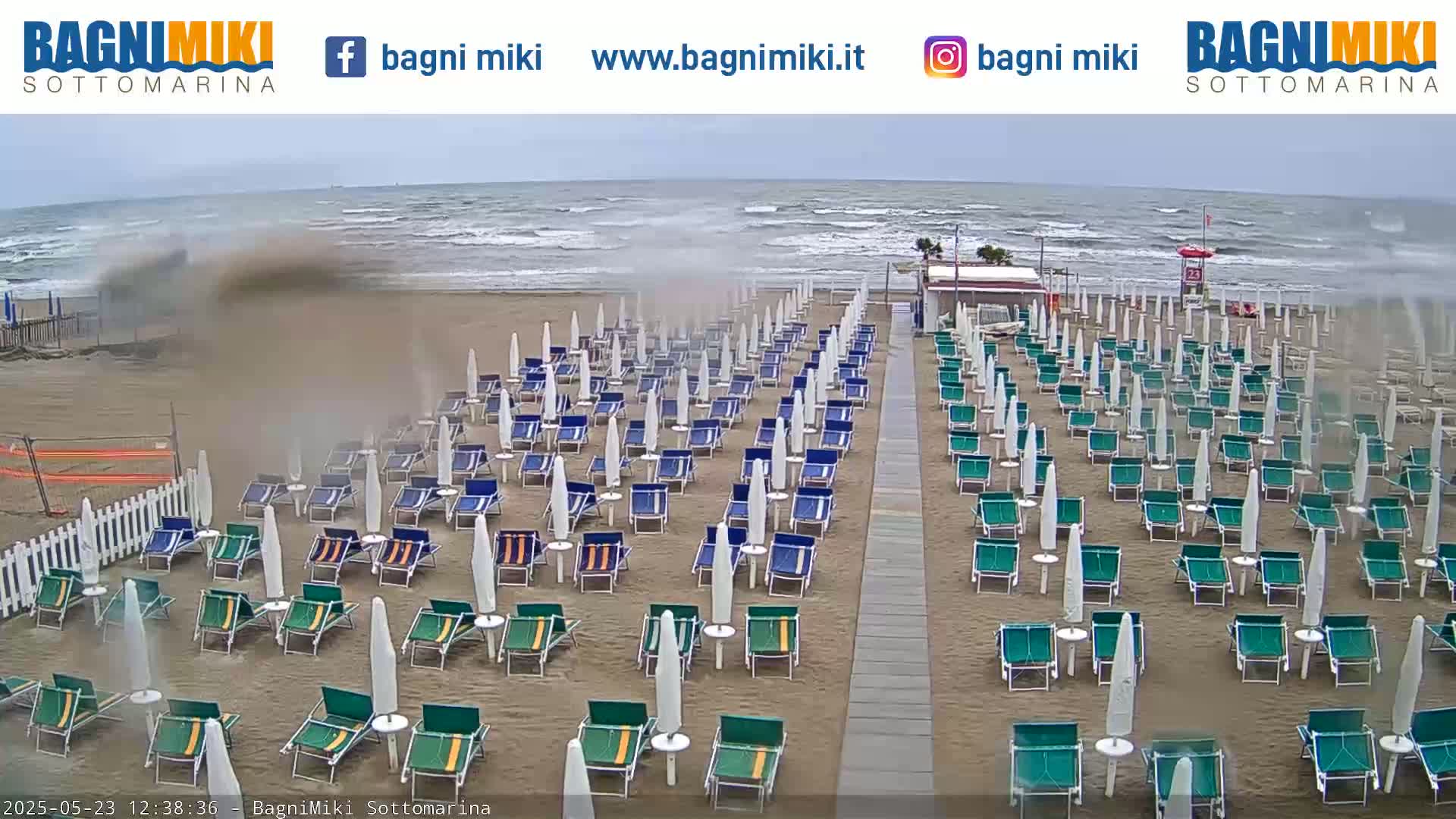 Rows of beach chairs and umbrellas are set up on a sandy beach with a rough sea under an overcast sky.
