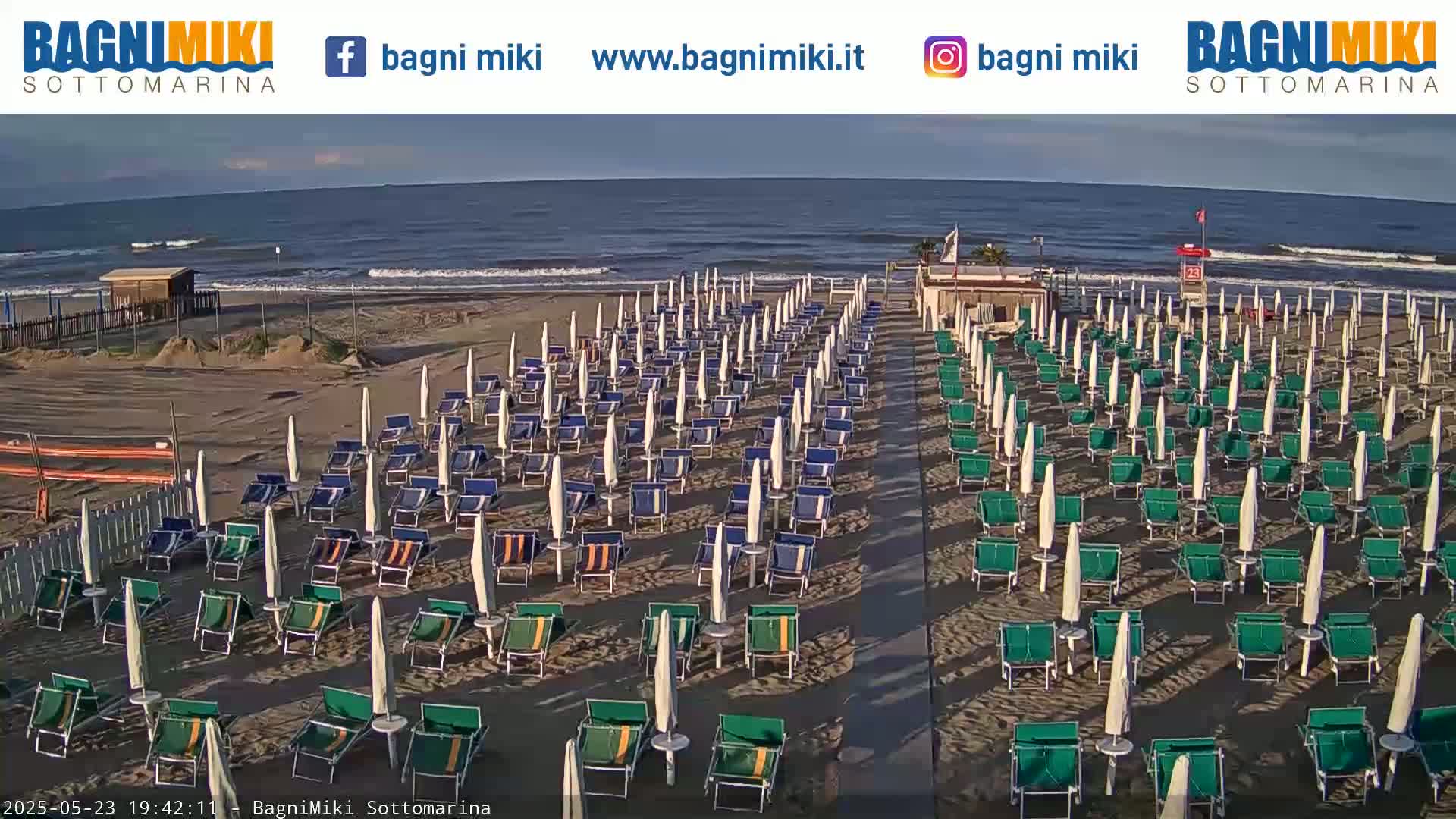 A beach with numerous rows of closed beach umbrellas and lounge chairs sits before a calm ocean under a clear sky.