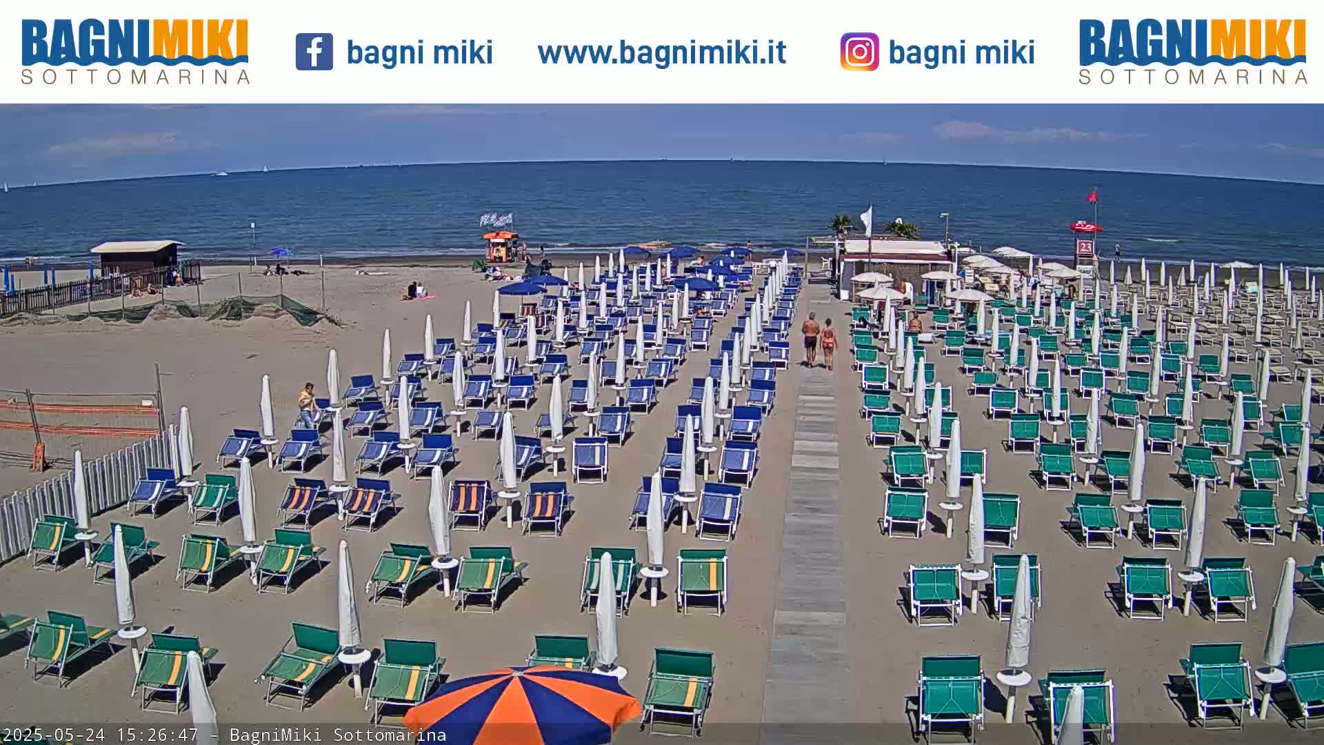 A sandy beach with numerous rows of green beach chairs and white umbrellas under a mostly clear sky, next to a calm ocean.
