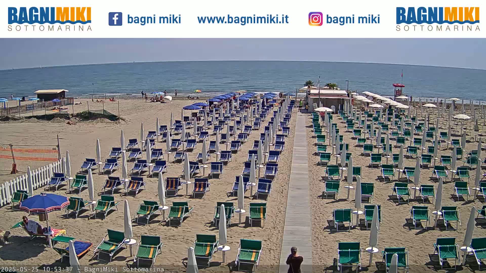 A sandy beach with numerous rows of umbrellas and lounge chairs under a clear, sunny sky, next to a calm ocean.