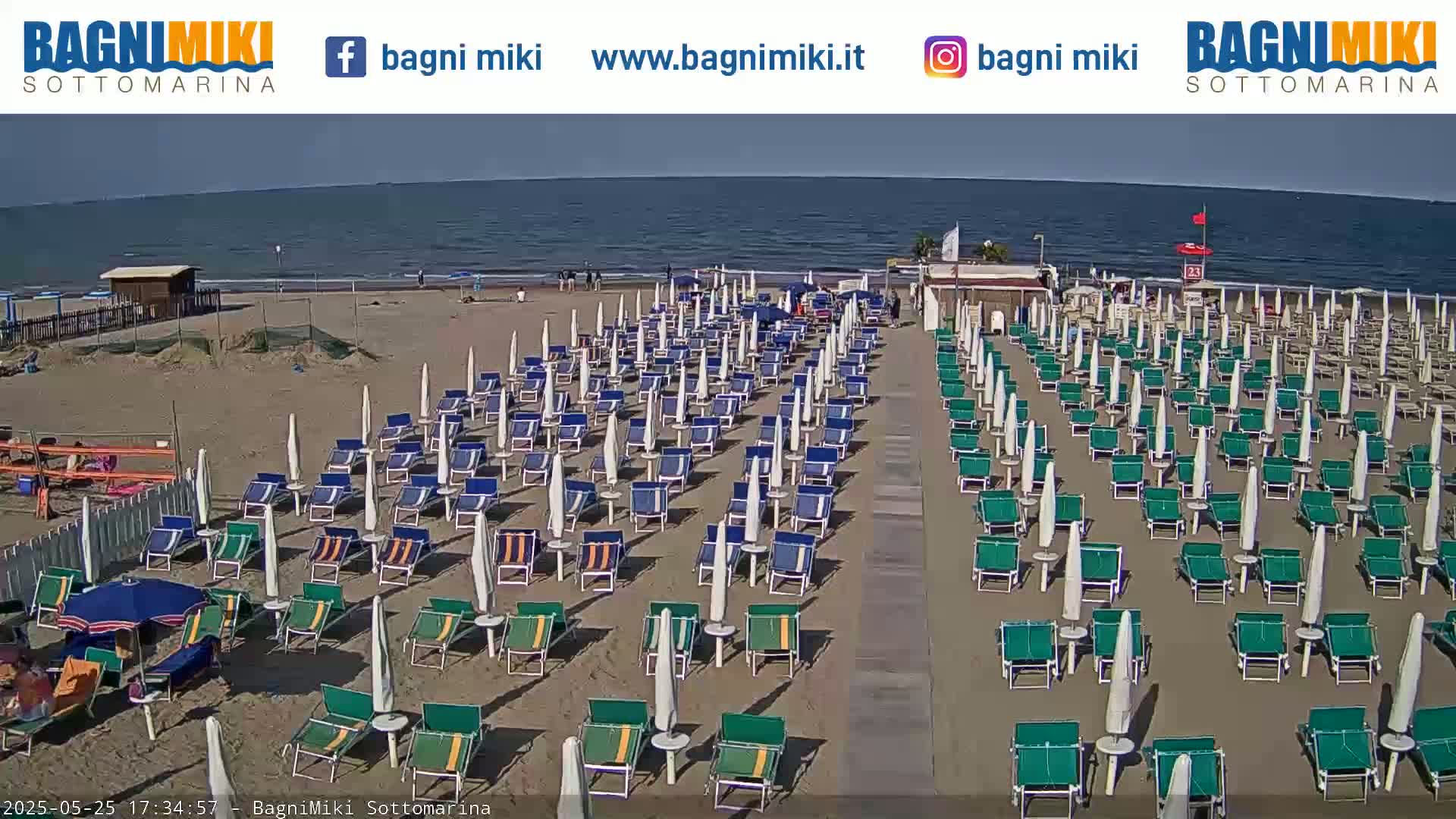 A sandy beach with numerous beach umbrellas and lounge chairs is shown on a sunny day, next to a calm sea.
