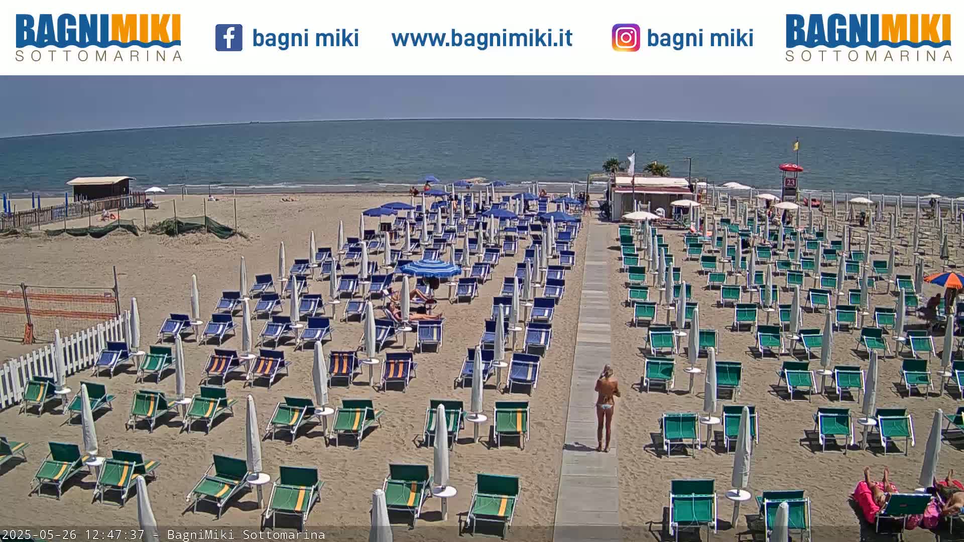 A sandy beach with numerous beach umbrellas and lounge chairs, under a sunny sky, overlooks a calm ocean.