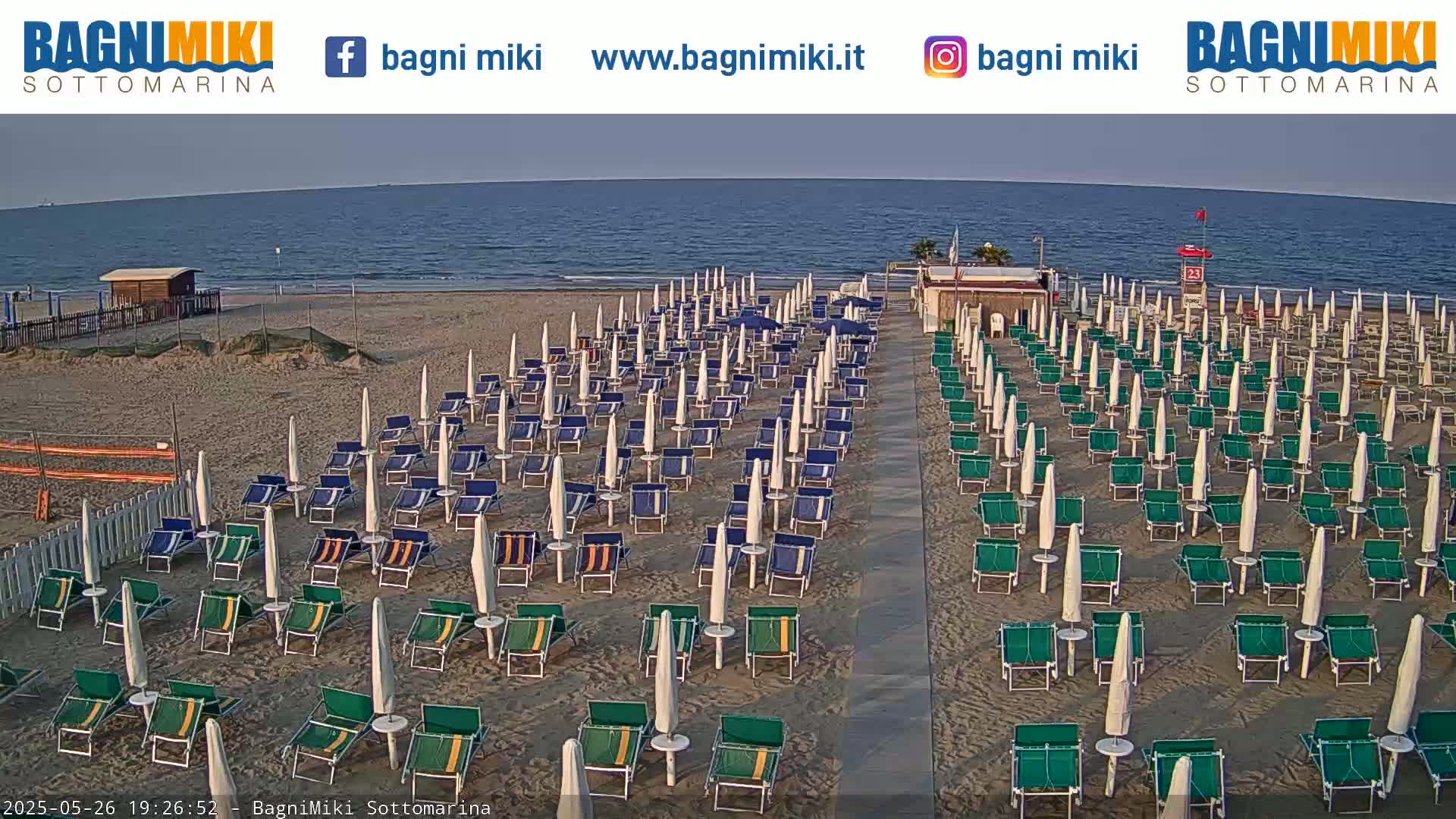 A sandy beach with numerous rows of closed beach umbrellas and lounge chairs is visible, overlooking a calm ocean under a clear sky.