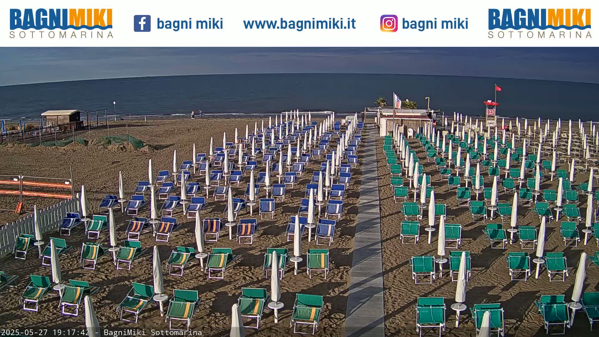 A sandy beach with numerous rows of closed beach umbrellas and lounge chairs sits before a calm ocean under a clear sky.