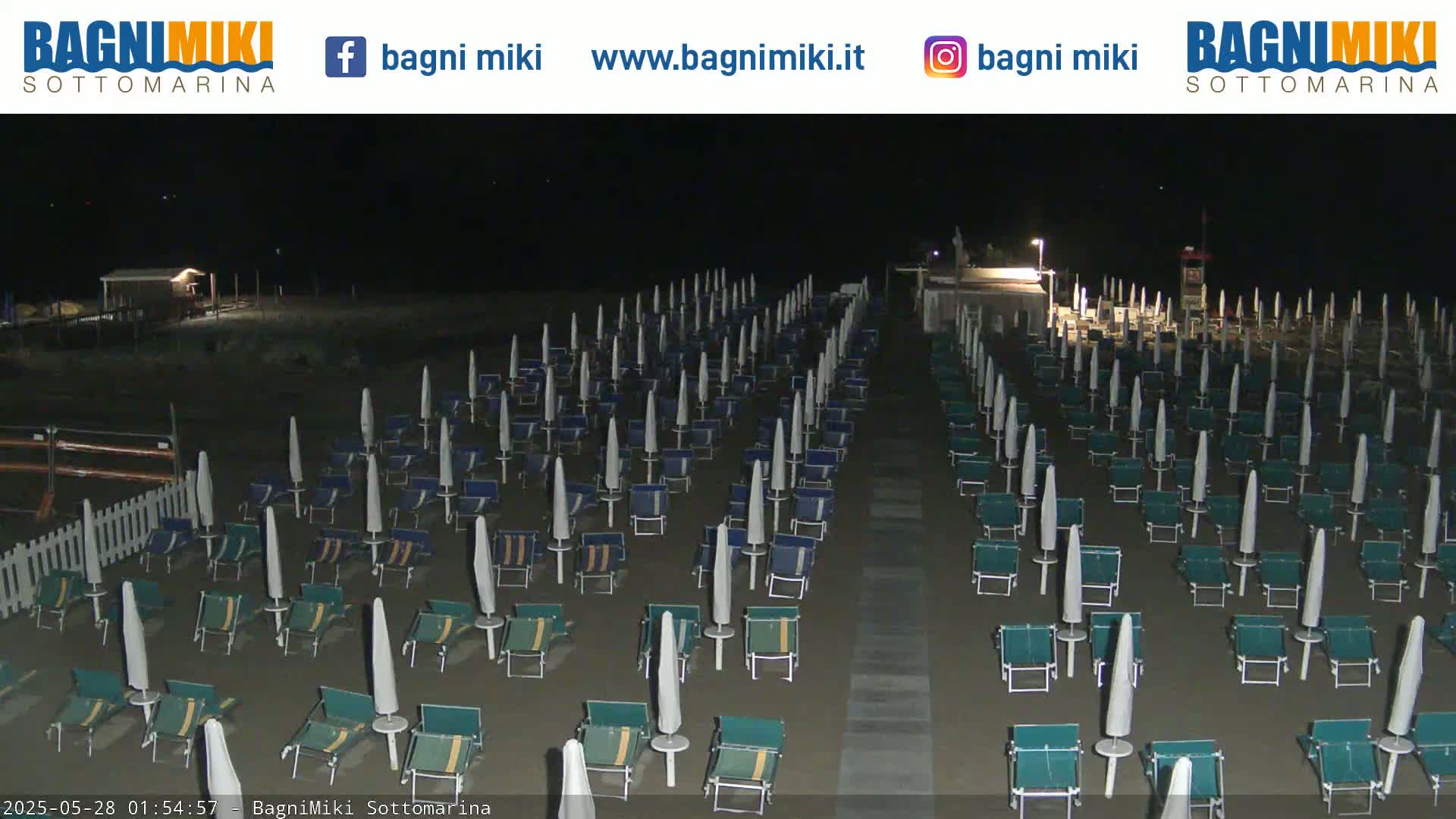 At night, a beach is shown with many closed beach umbrellas and lounge chairs arranged in rows on dark sand.