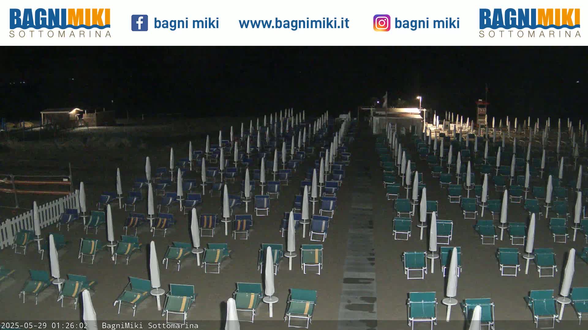At night, a sandy beach is lined with rows of closed beach umbrellas and chairs under artificial lighting.