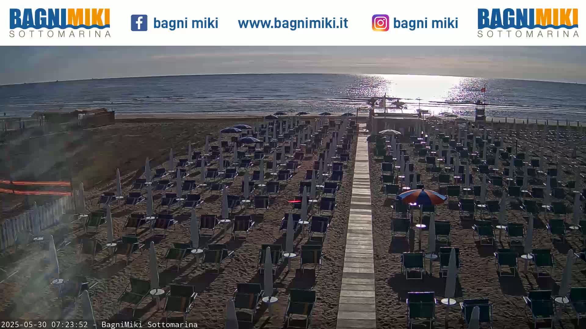 A sandy beach with rows of beach chairs and umbrellas under a partly sunny sky, next to a calm ocean.