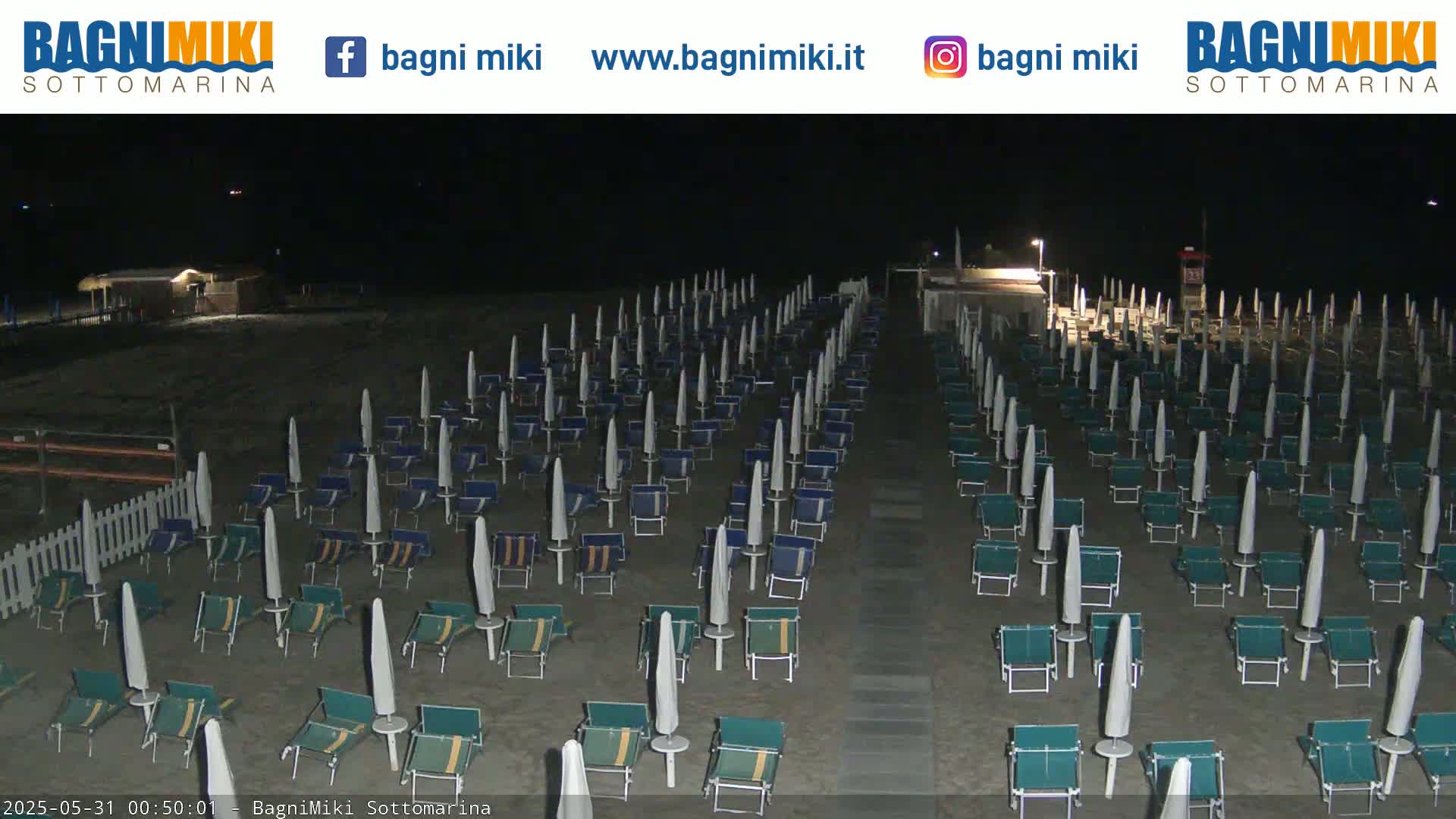 A beach at night is shown with many closed beach umbrellas and chairs arranged in rows under artificial lights.