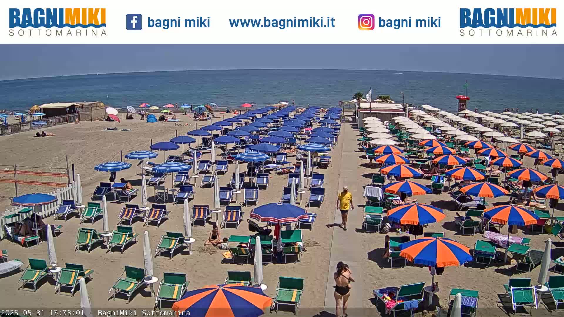 A sandy beach with numerous blue and orange umbrellas and beach chairs under a sunny sky, next to a calm blue ocean.
