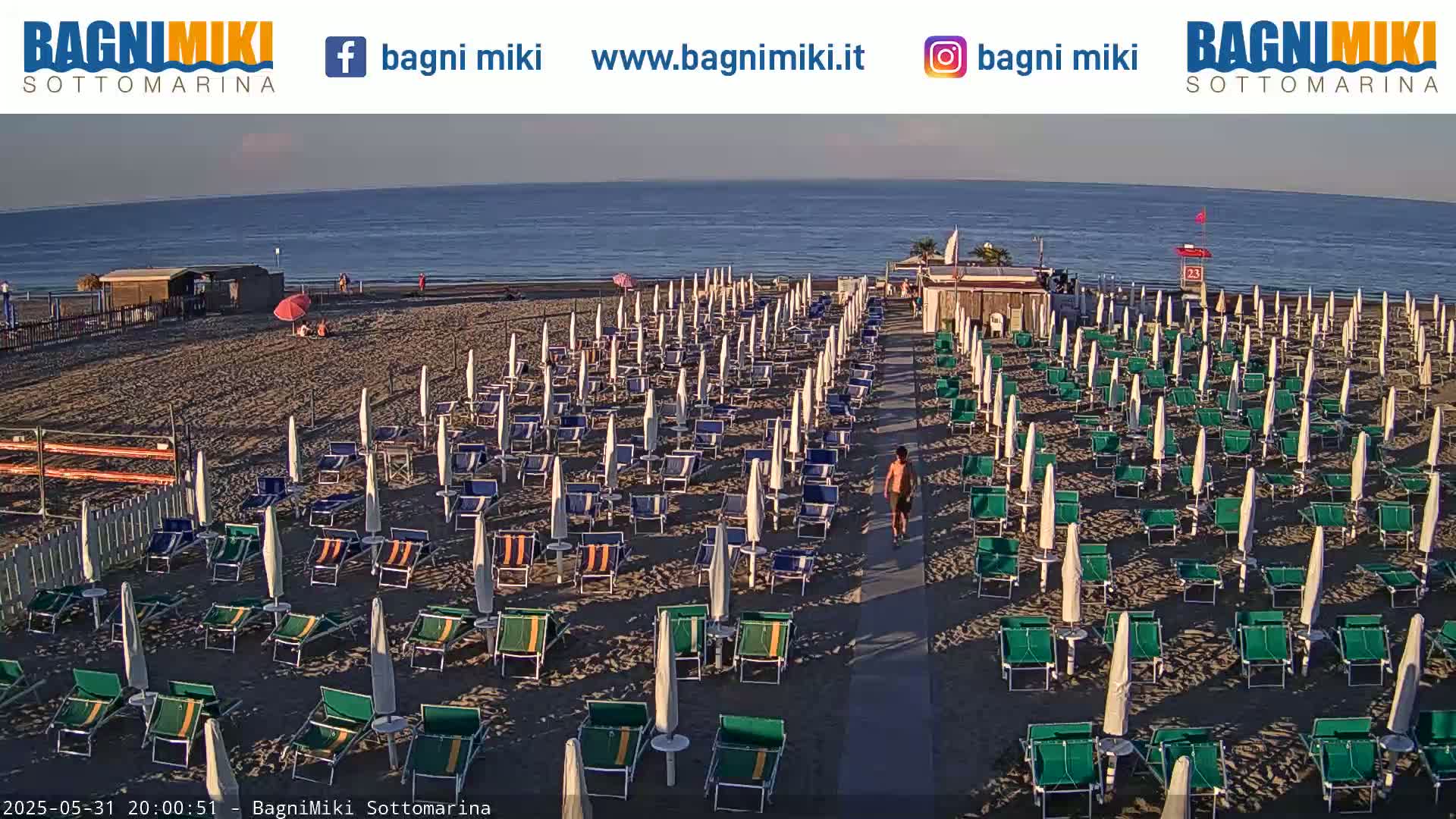 A mostly empty beach with rows of closed beach umbrellas and lounge chairs under a clear evening sky, overlooking a calm sea.