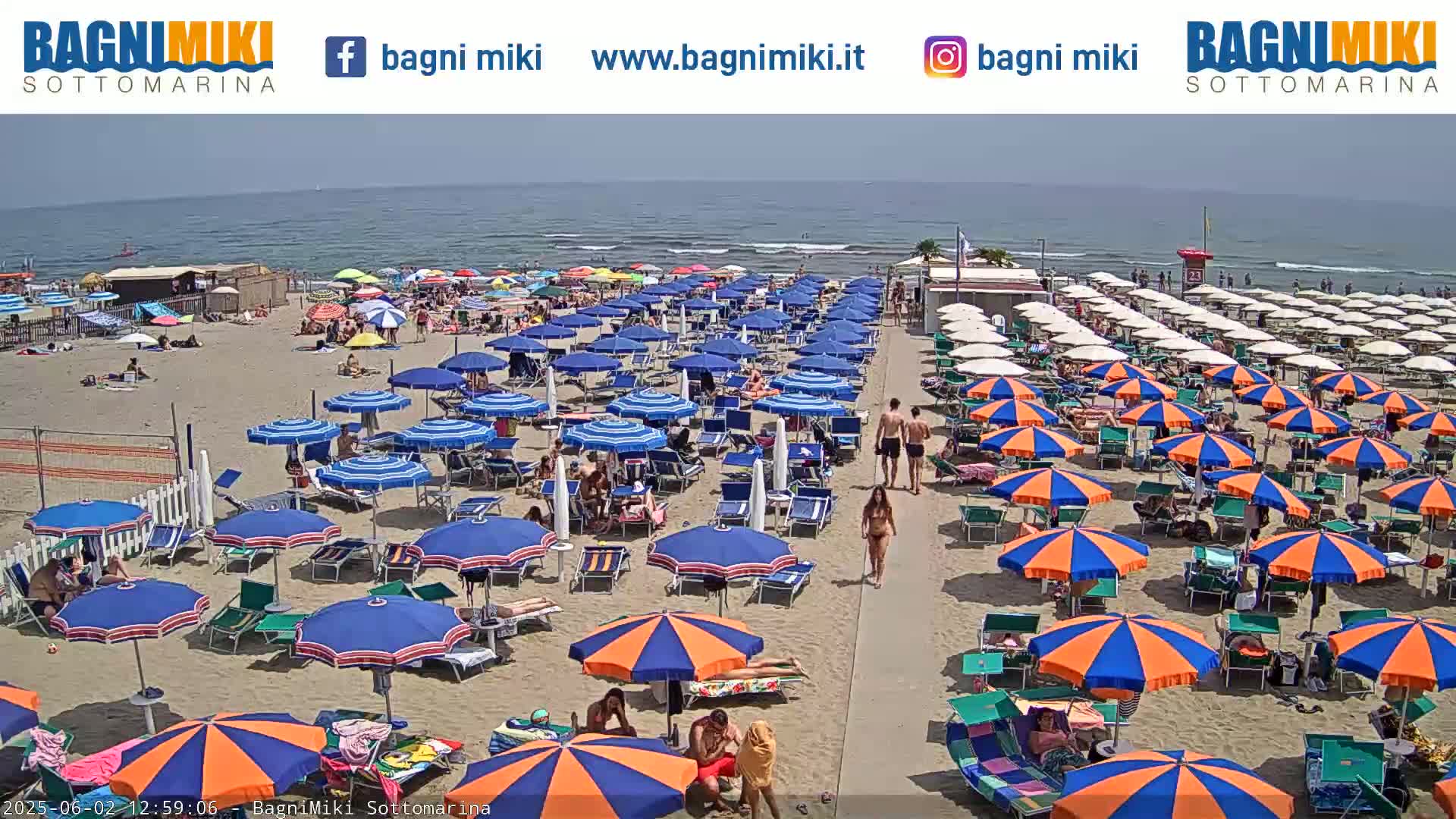 A sandy beach is crowded with numerous blue and orange umbrellas and beachgoers on a sunny day.