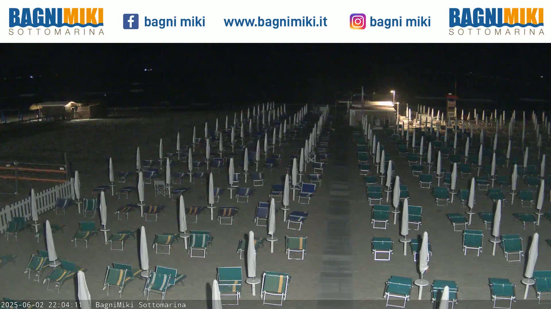 A dark beach at night is shown with numerous closed beach umbrellas and lounge chairs arranged in rows.