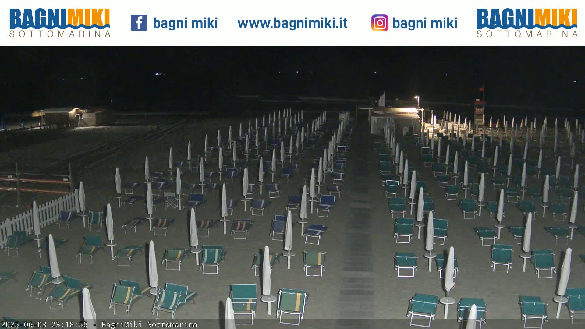 Rows of closed beach umbrellas and lounge chairs stand on a dark sandy beach at night.