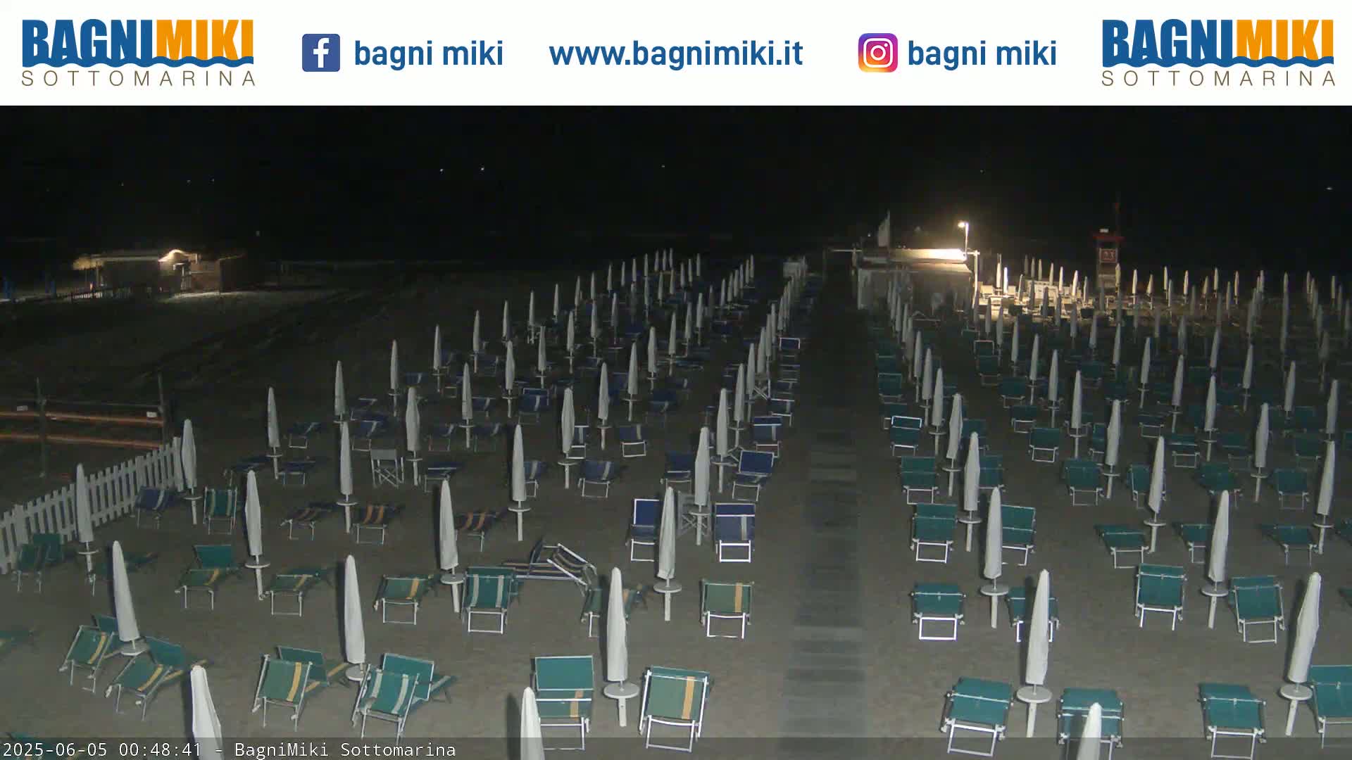 A beach at night is shown with many closed beach umbrellas and lounge chairs arranged in rows under dark night skies.
