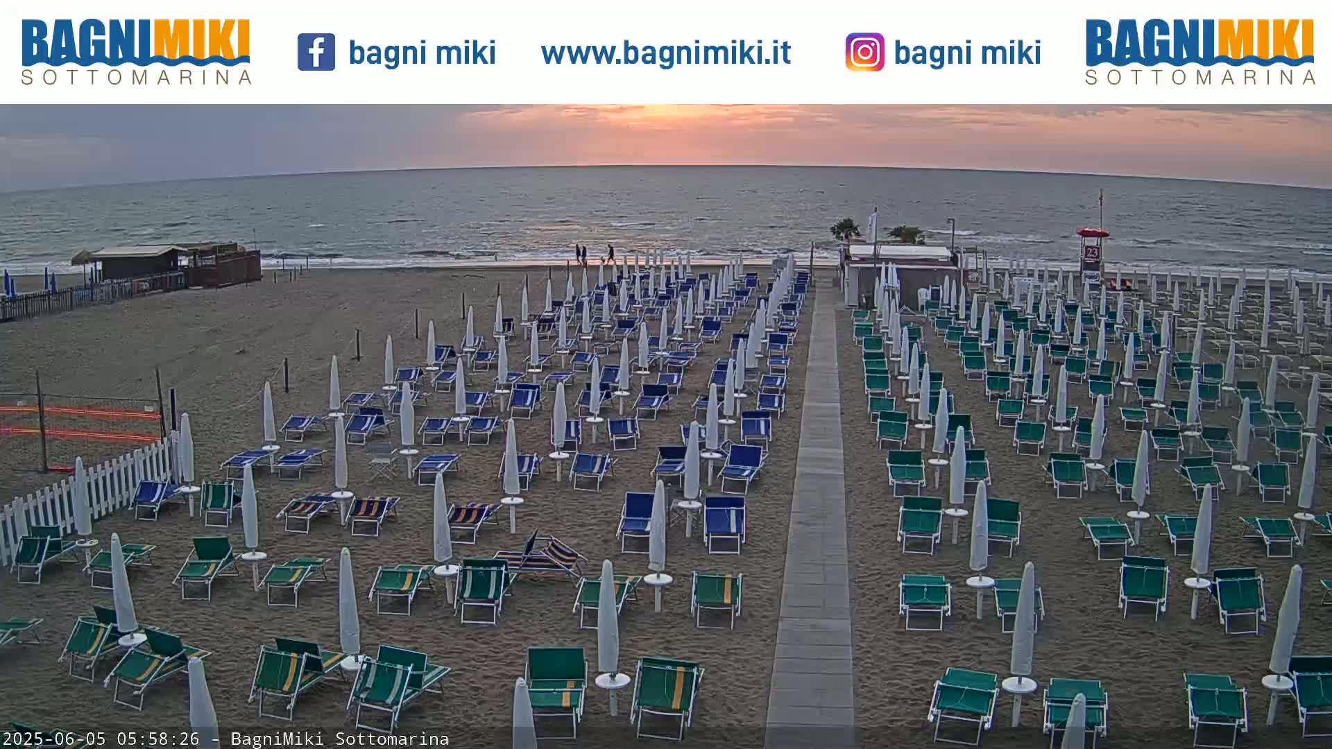 A sandy beach with numerous rows of closed beach umbrellas and lounge chairs is shown at sunrise, with calm ocean waves visible in the background.