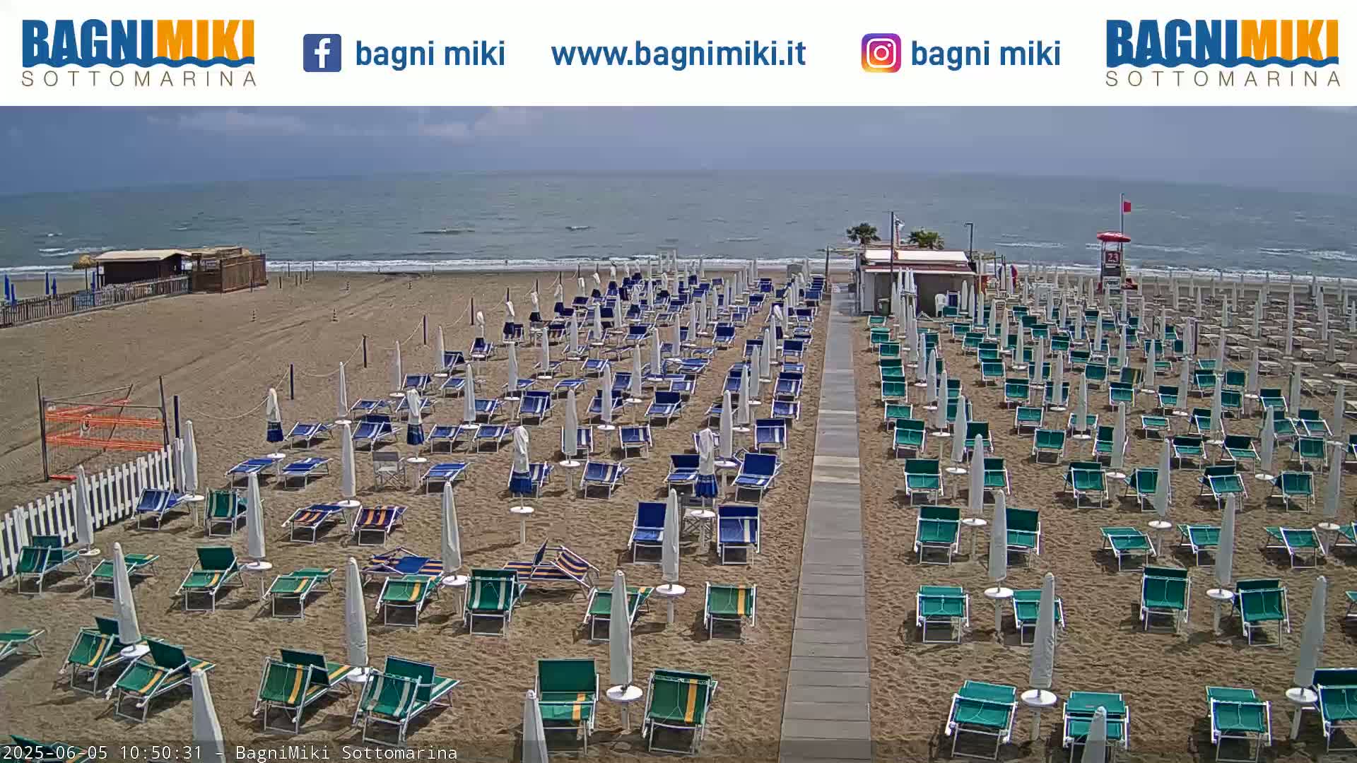 A mostly sunny beach scene shows rows of empty beach chairs and umbrellas on a sandy beach next to a calm ocean.