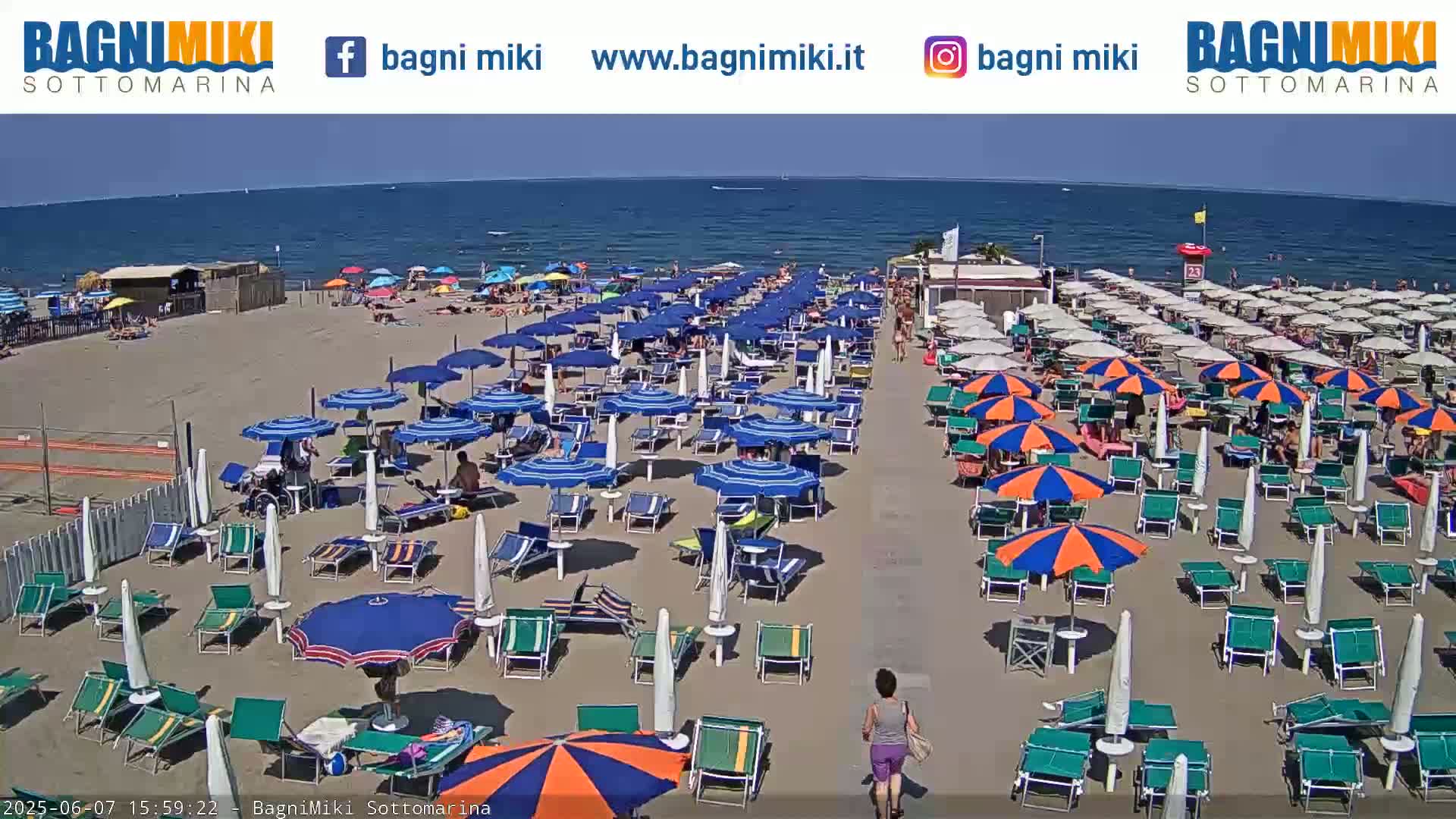A sandy beach with numerous blue and orange umbrellas and lounge chairs is situated next to a calm blue ocean under a sunny sky.