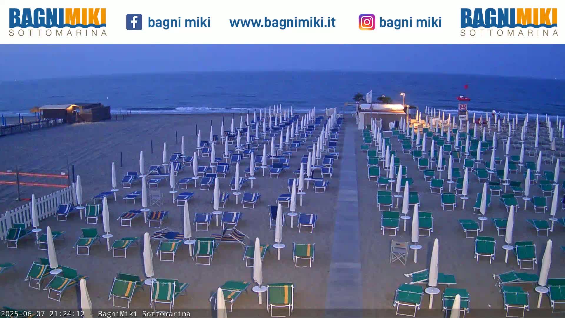 Rows of beach umbrellas and lounge chairs are set up on a sandy beach next to a calm, dark-blue sea under a twilight sky.