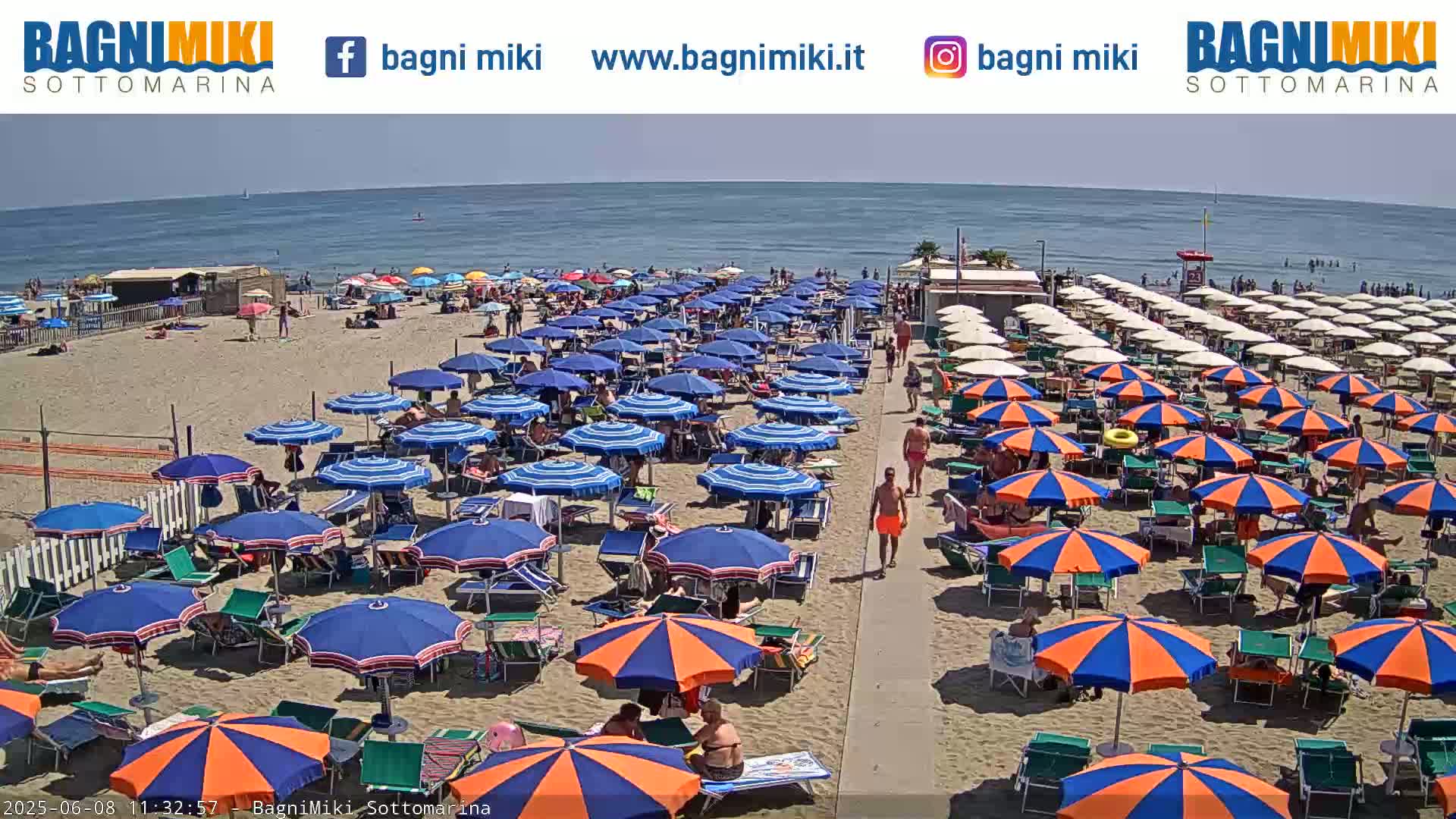 A sunny beach scene shows many blue and orange umbrellas and beach chairs on a sandy beach next to a calm ocean.