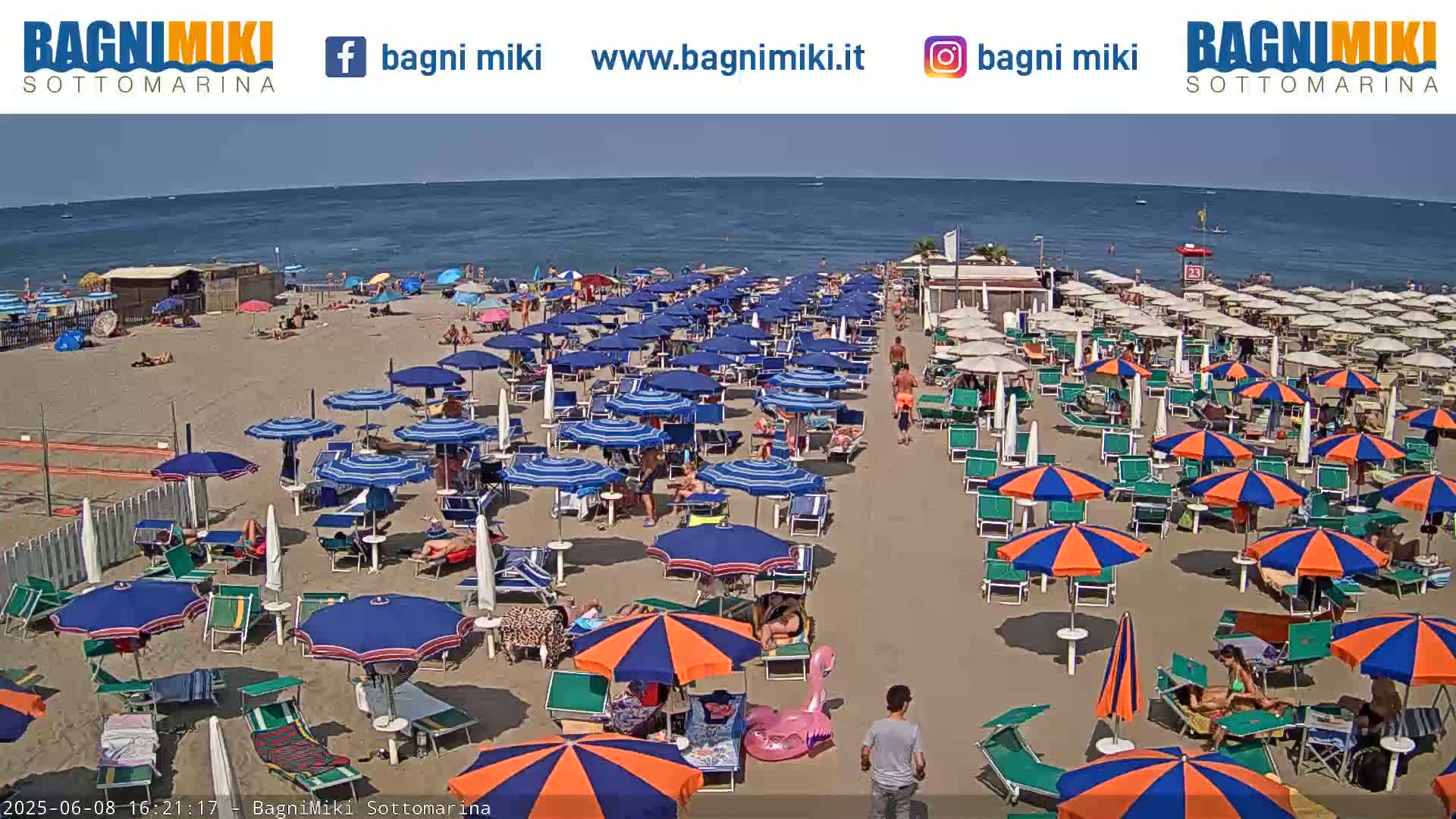 A sandy beach with numerous blue and orange umbrellas and beach chairs, under a sunny sky, next to a calm sea.