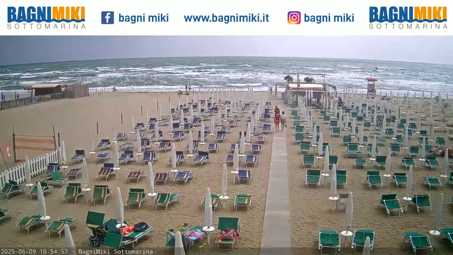 A mostly empty beach with many closed umbrellas and lounge chairs sits under an overcast sky with a wavy ocean in the background.