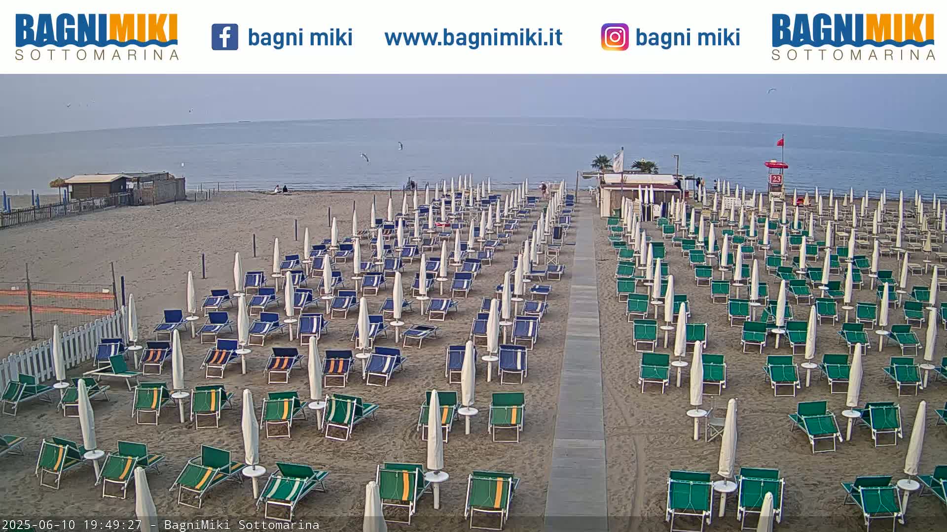 A sandy beach with numerous rows of closed beach umbrellas and chairs is visible, overlooking a calm sea under a mostly clear sky.