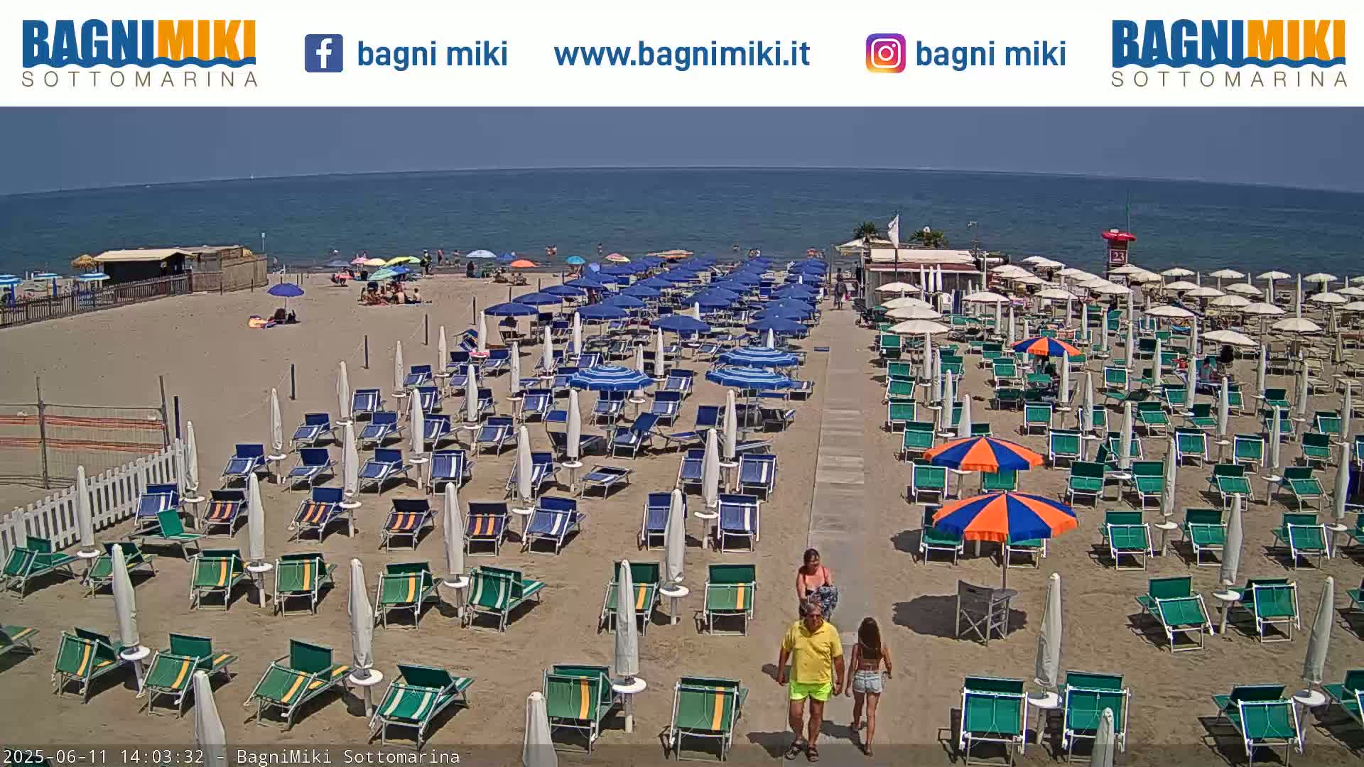 A sandy beach with numerous rows of beach umbrellas and lounge chairs is shown on a sunny day, next to a calm ocean.