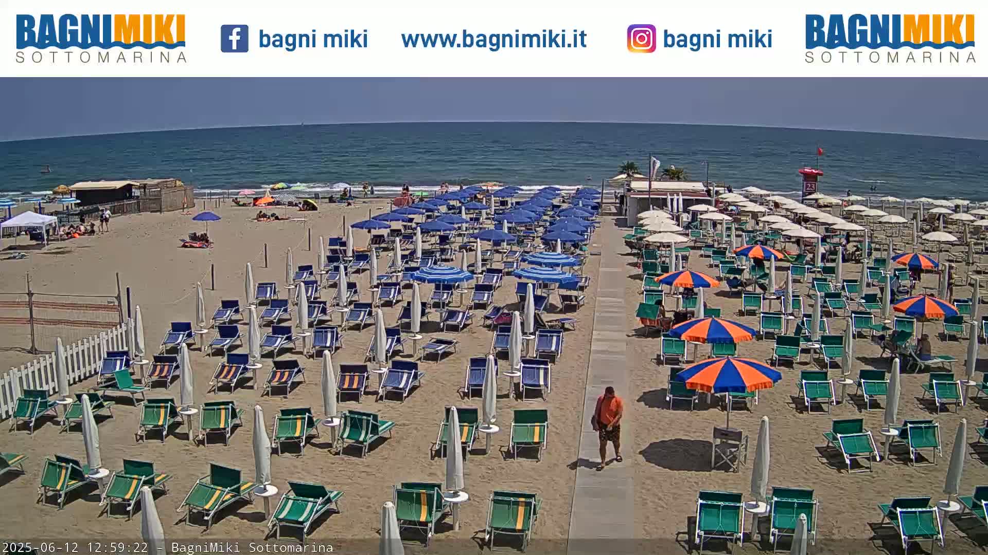 A sandy beach with numerous beach umbrellas and lounge chairs is shown on a sunny day next to a calm ocean.
